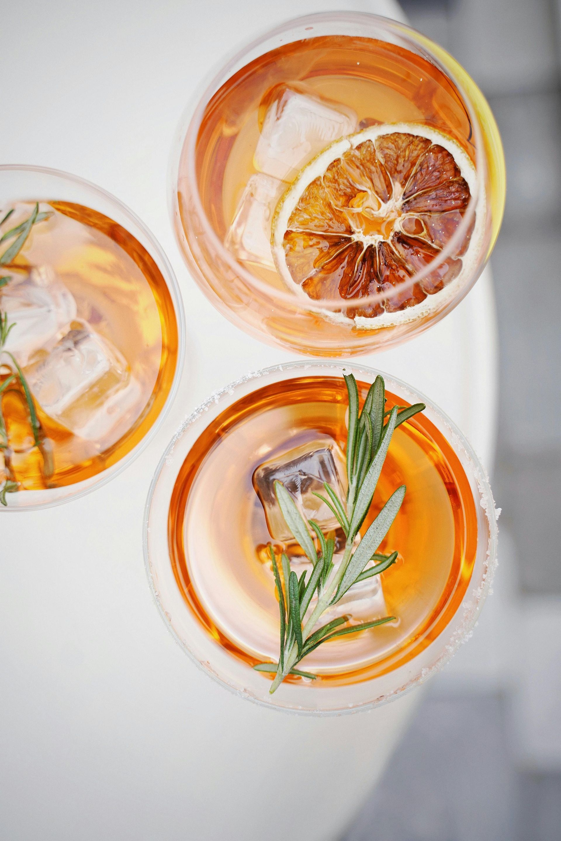 Three glasses of orange juice with ice cubes and rosemary on a table.