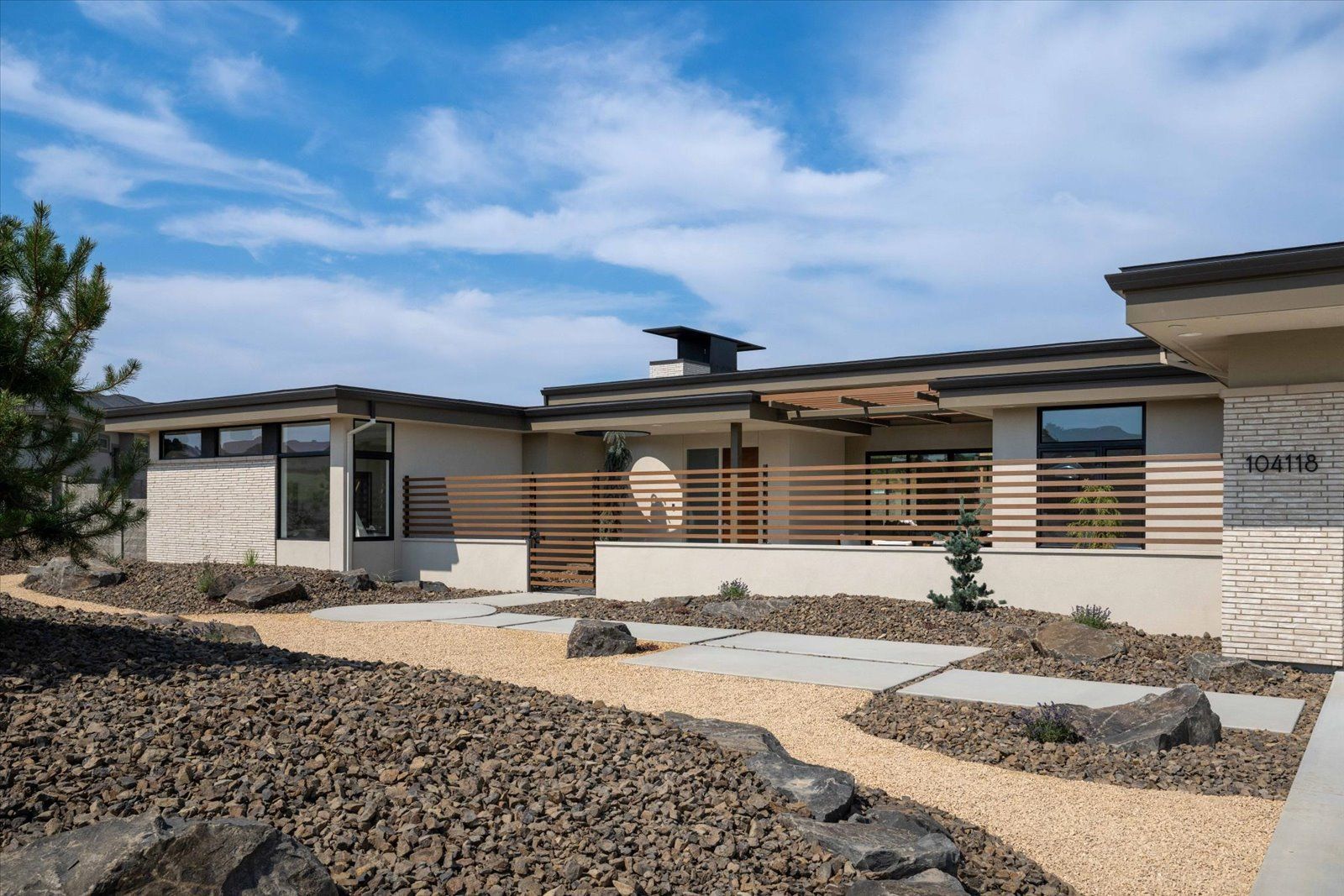 Modern home with tan facade, stone accents, wooden fencing, and xeriscaping under a blue sky.