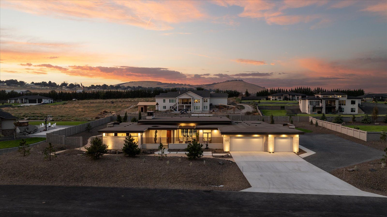 Modern home with three-car garage and long driveway, set against a sunset, and rolling hills.