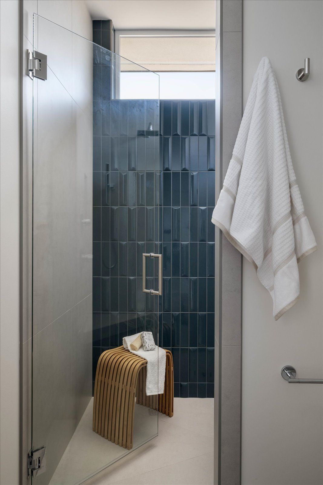 Modern bathroom with blue tile shower, glass door, and wooden stool. White towel hangs on the door.