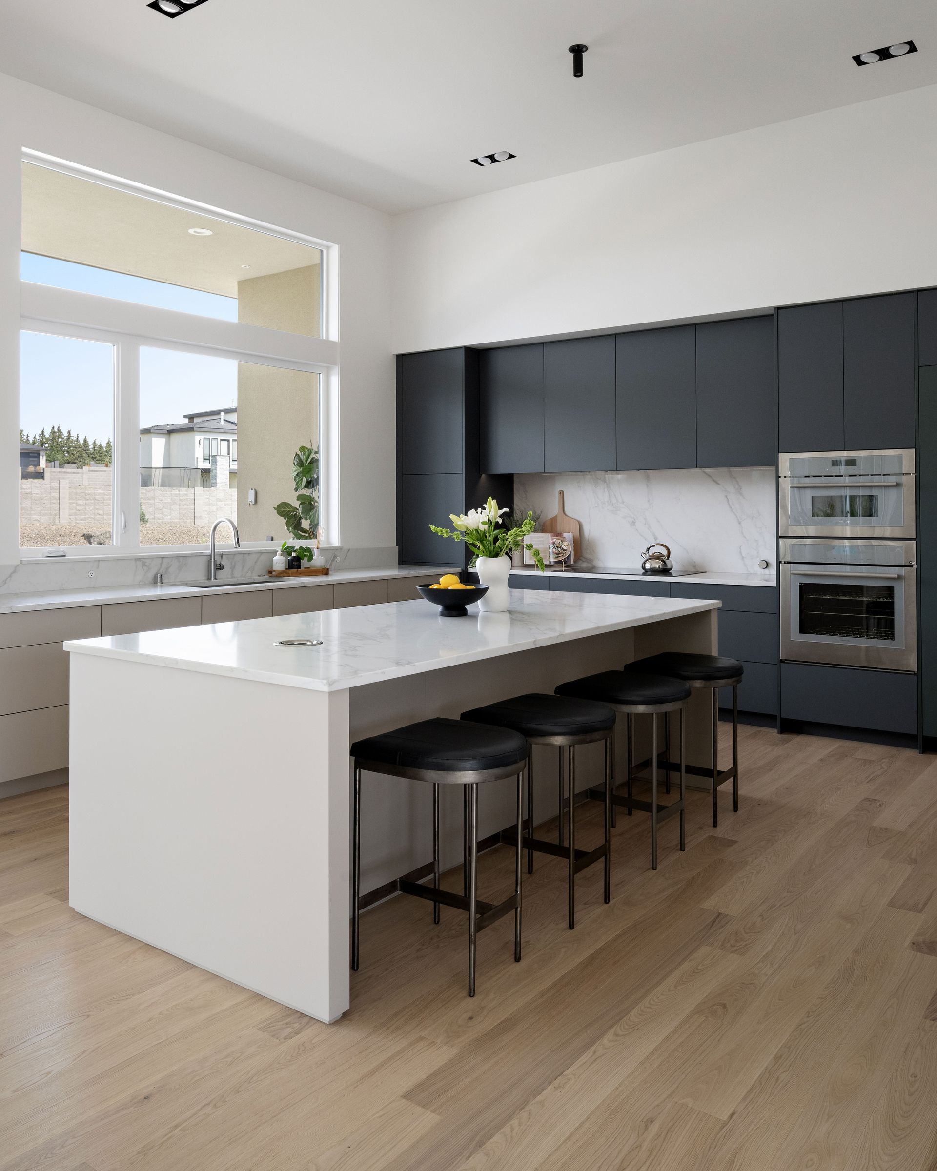 Modern kitchen with island, dark cabinets, and large window. Wood floors, stools, and white countertop.