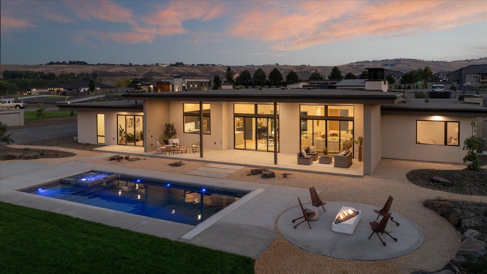 Modern home with pool and fire pit at dusk. Exterior with large windows, landscaping, and a view of the hills.