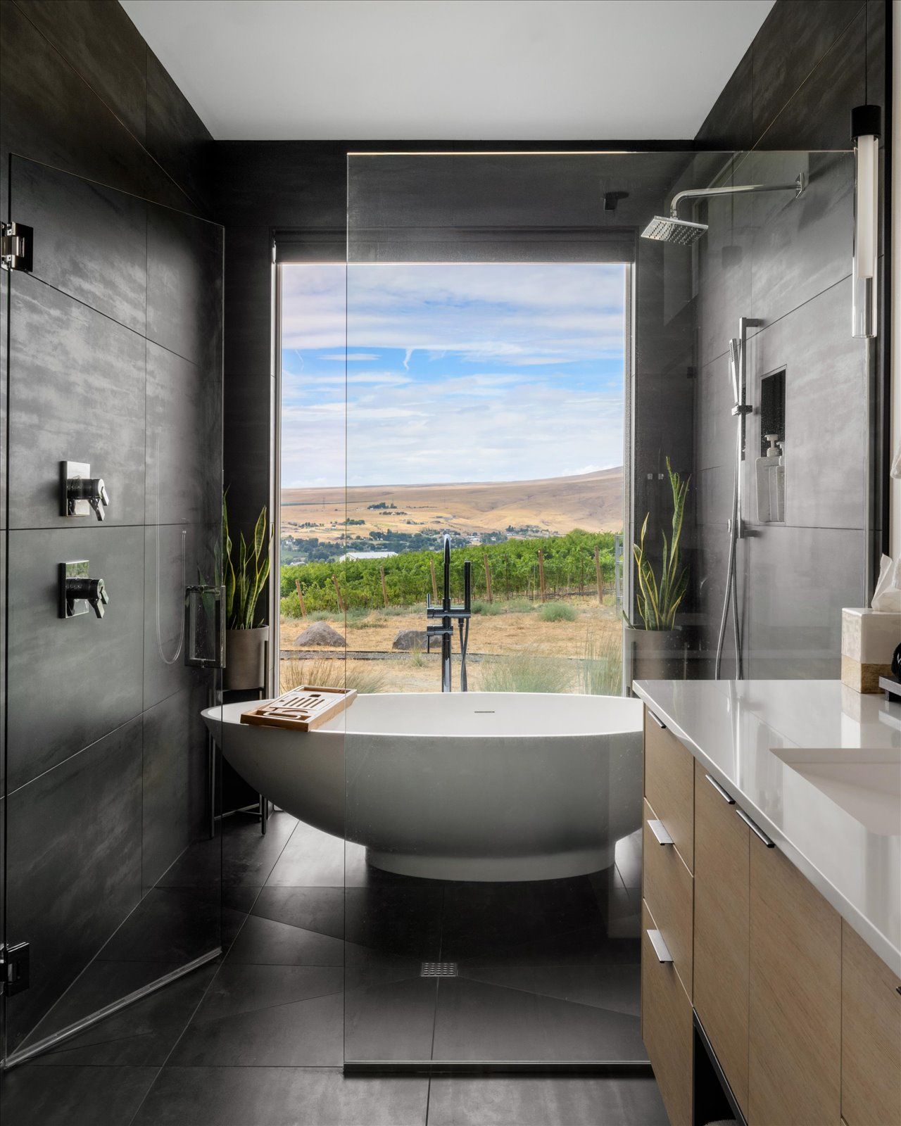 Modern bathroom with dark tile, large window overlooking a vineyard, and a white freestanding bathtub.