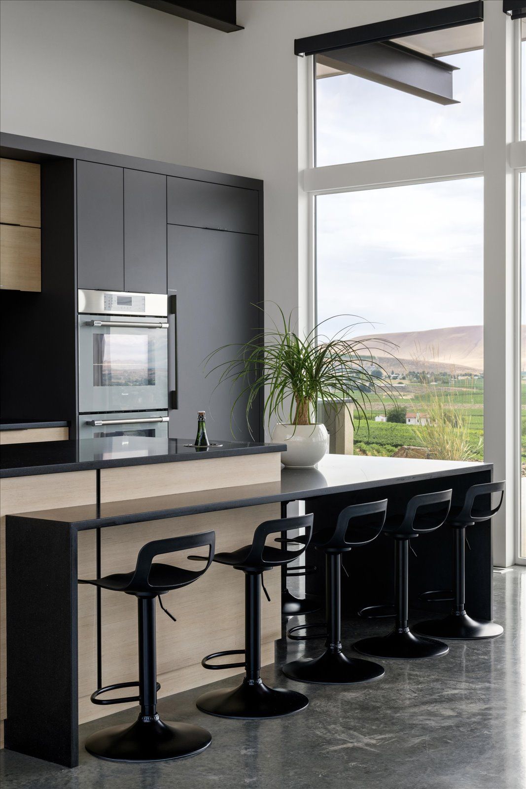 Modern kitchen with black cabinets and island, bar stools, and large window overlooking a landscape.