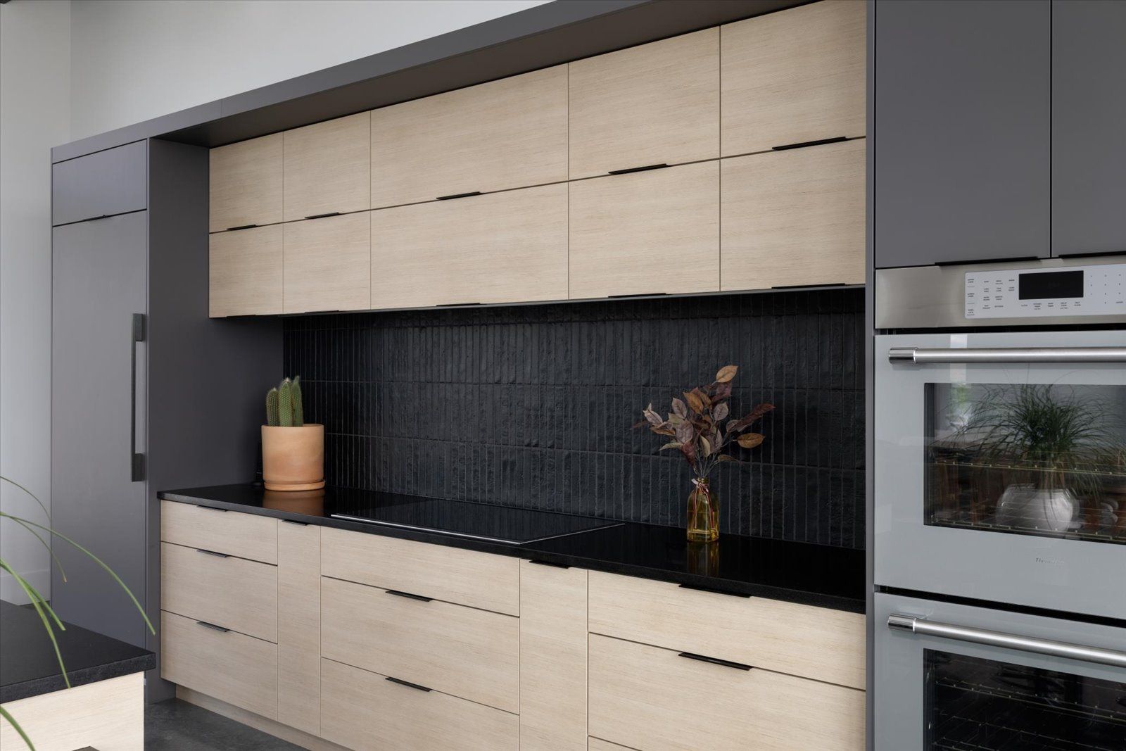 Modern kitchen with light wood and gray cabinets, black backsplash, built-in ovens.