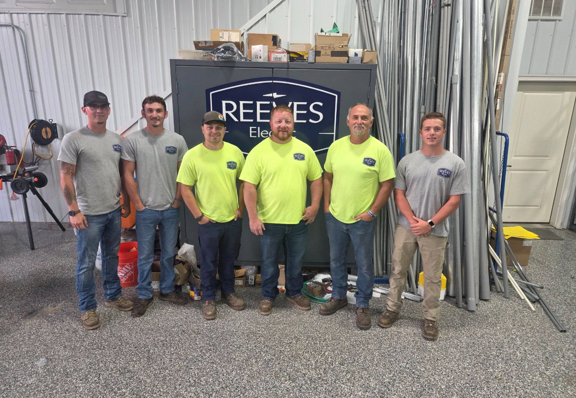 Six men posing in front of a company logo. They wear work clothes in a workshop setting. Six men posing in front of a company logo. They wear work clothes in a workshop setting.