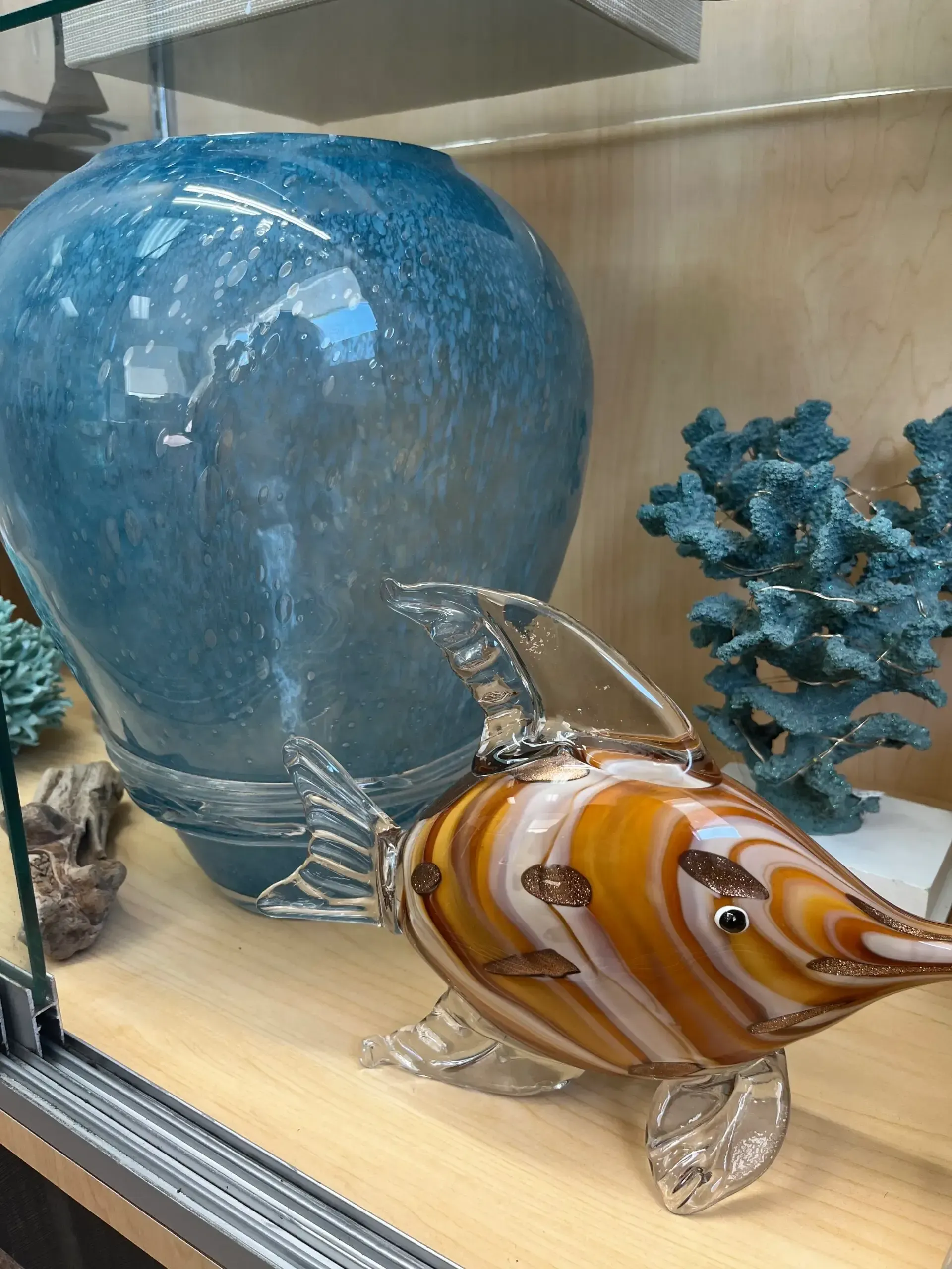 A glass fish sculpture with orange and white swirls sits next to a large blue vase and a faux blue coral on a shelf.