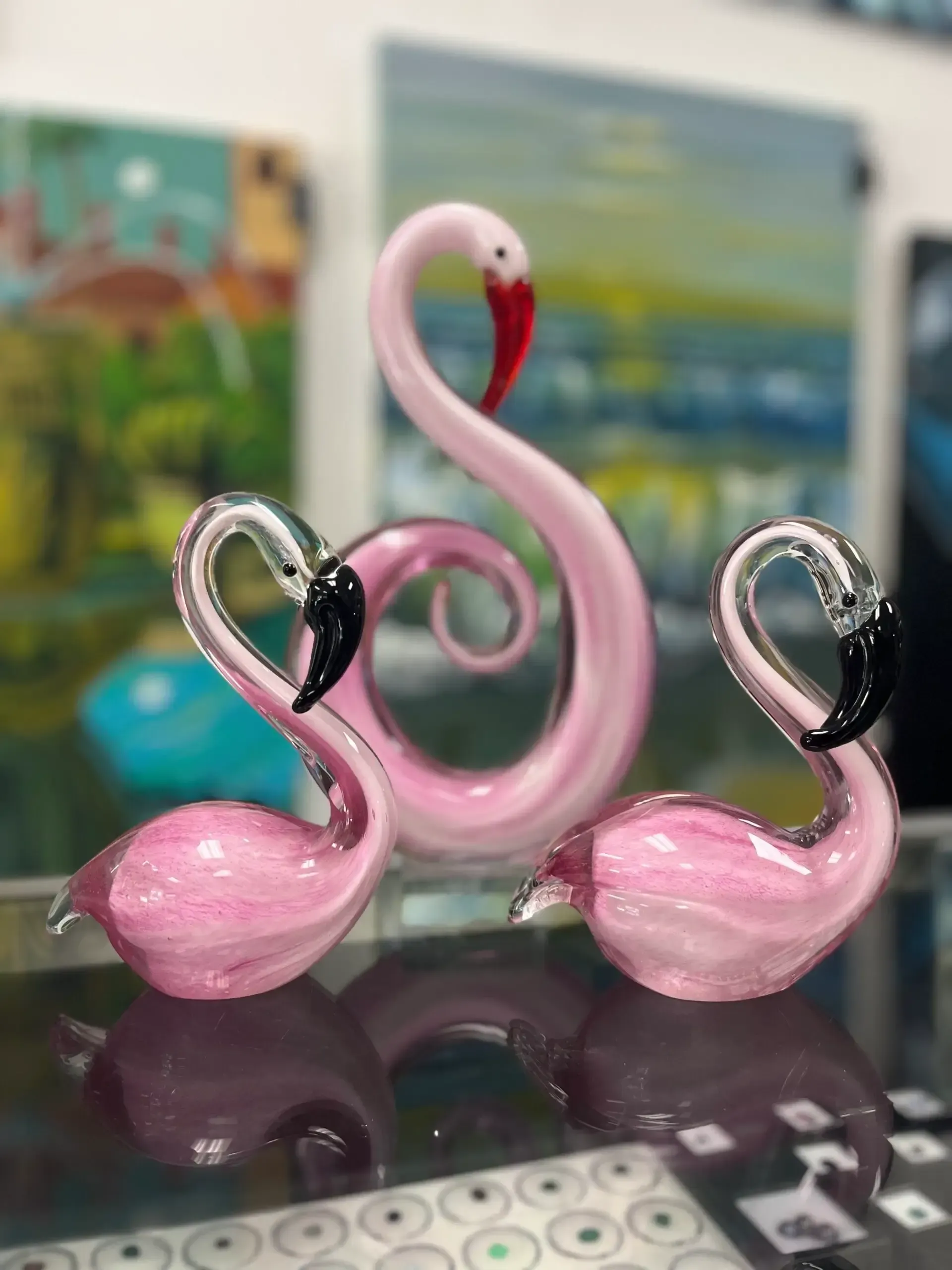 Three glass flamingo figurines, one standing tall in the back, with two smaller ones in front on a reflective surface.