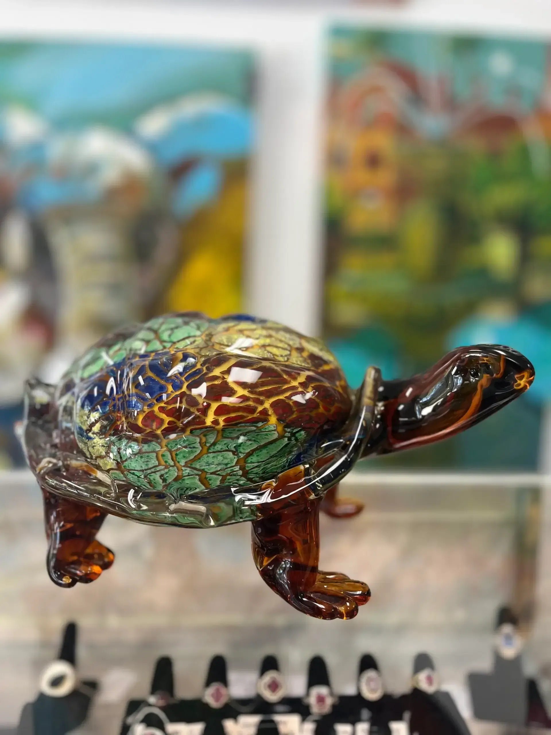 A decorative glass turtle with a mosaic-patterned shell in shades of brown, green, and blue, perched on a display shelf.