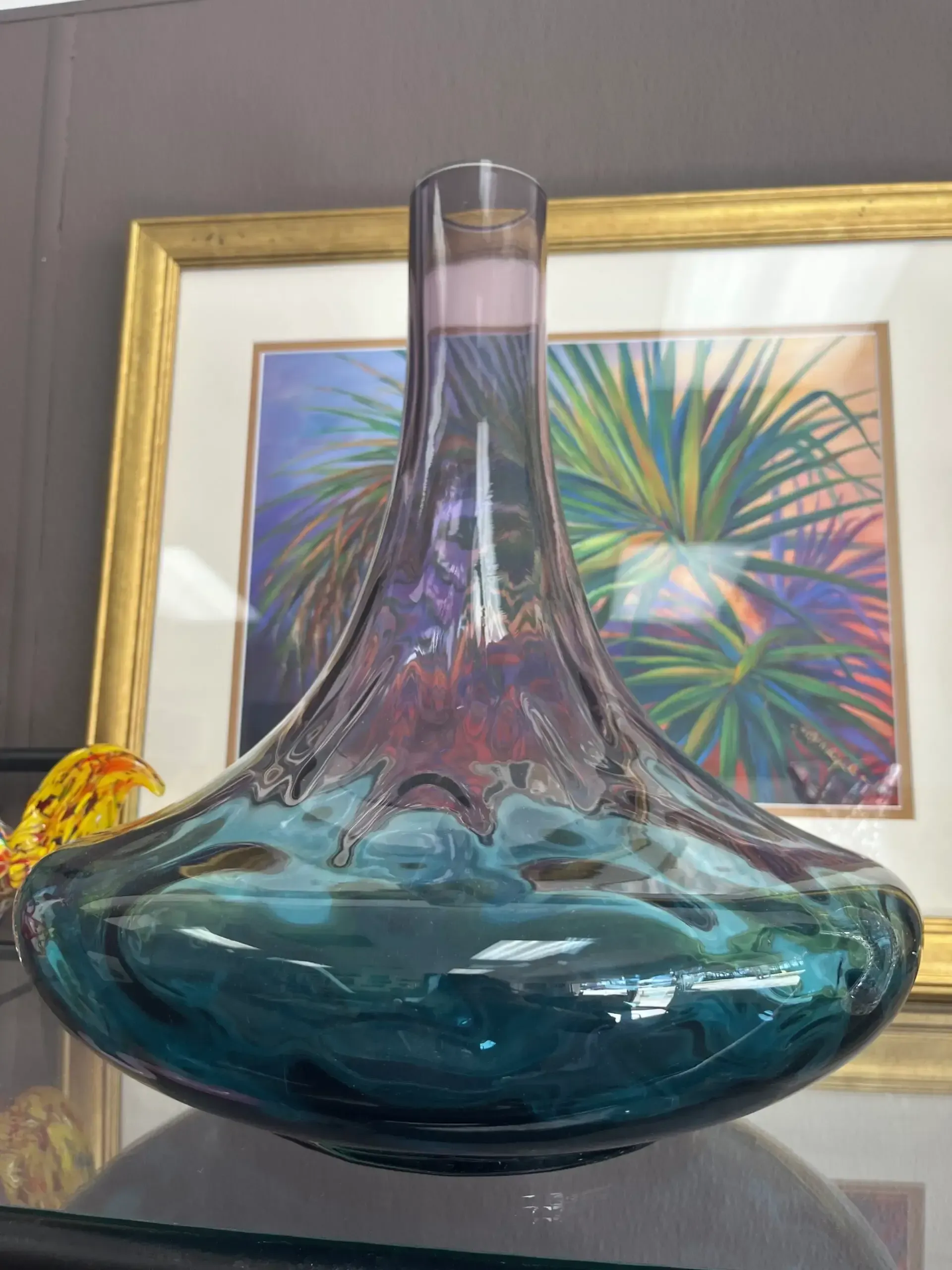 A wide-bottomed glass decanter with a teal base fading into a tall, purple neck, sitting in front of a framed painting.