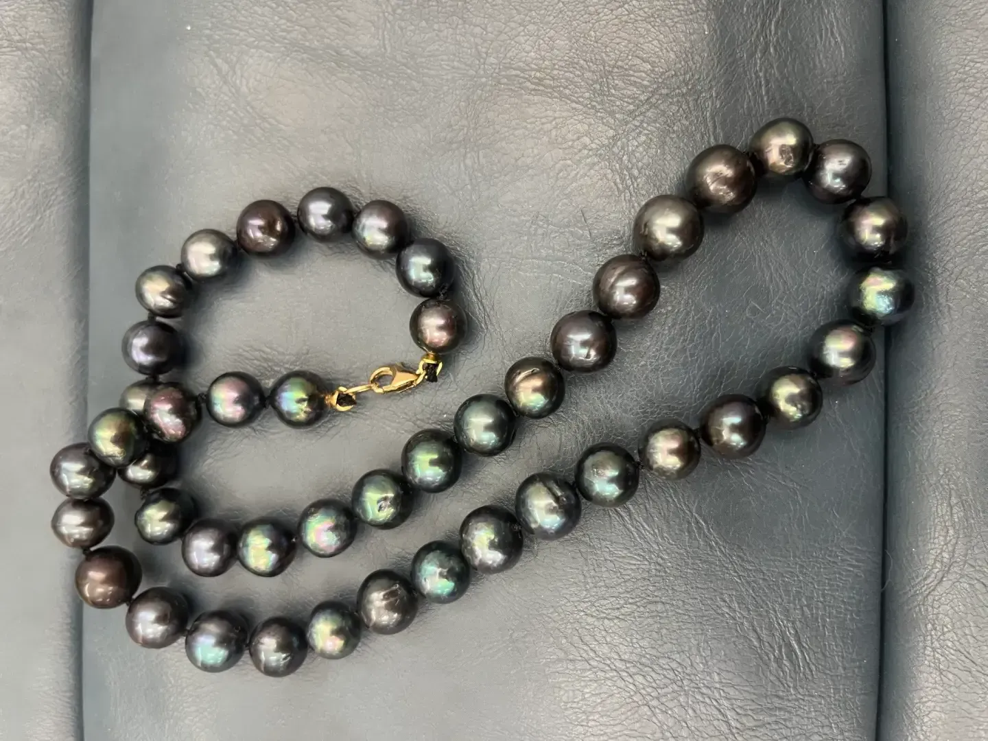 A strand of lustrous, dark gray or black pearls with iridescent undertones and a gold clasp, resting on a gray surface.