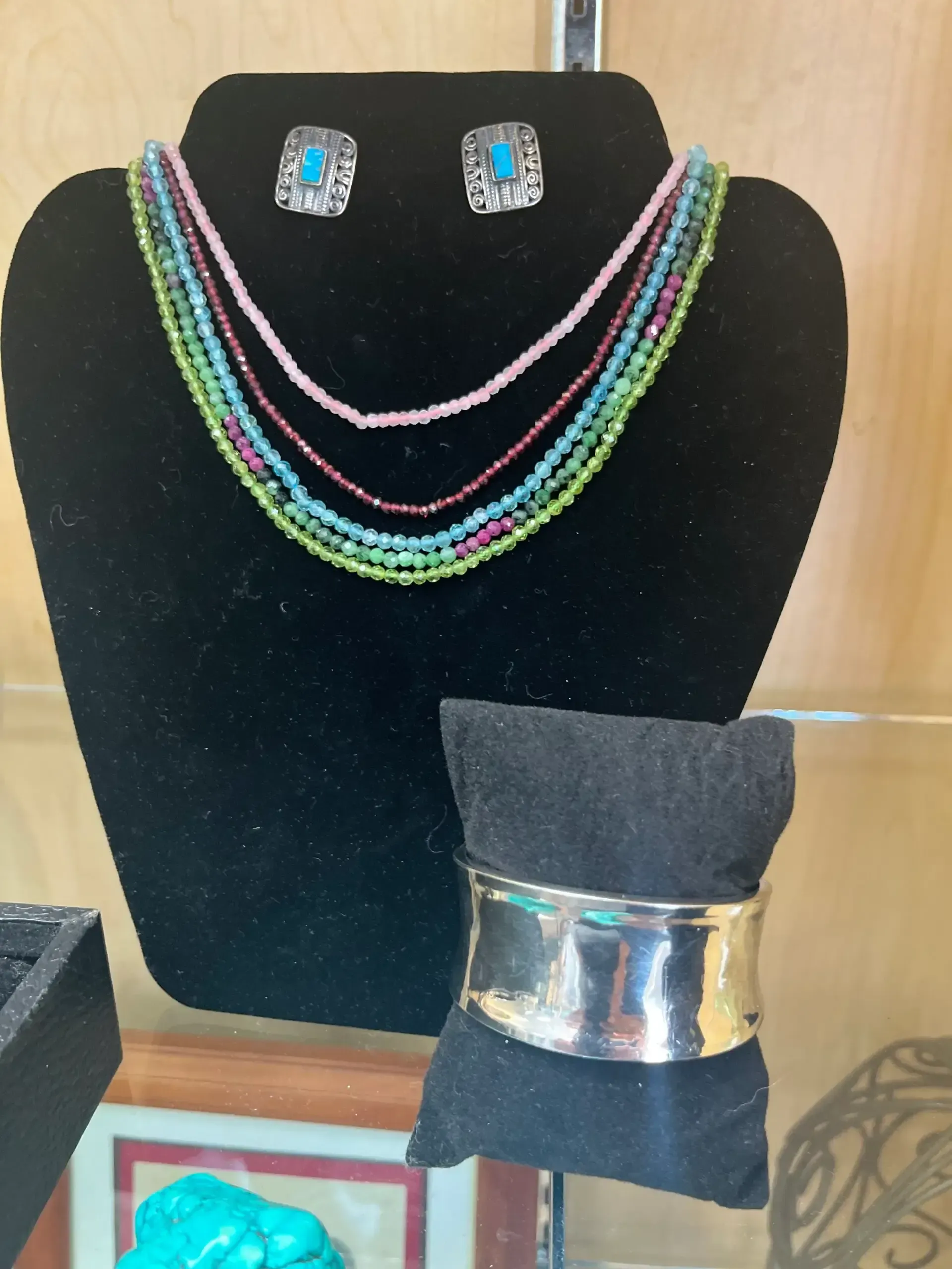 A tiered multi-colored beaded necklace, matching stud earrings, and a wide silver cuff bracelet on a black display stand.