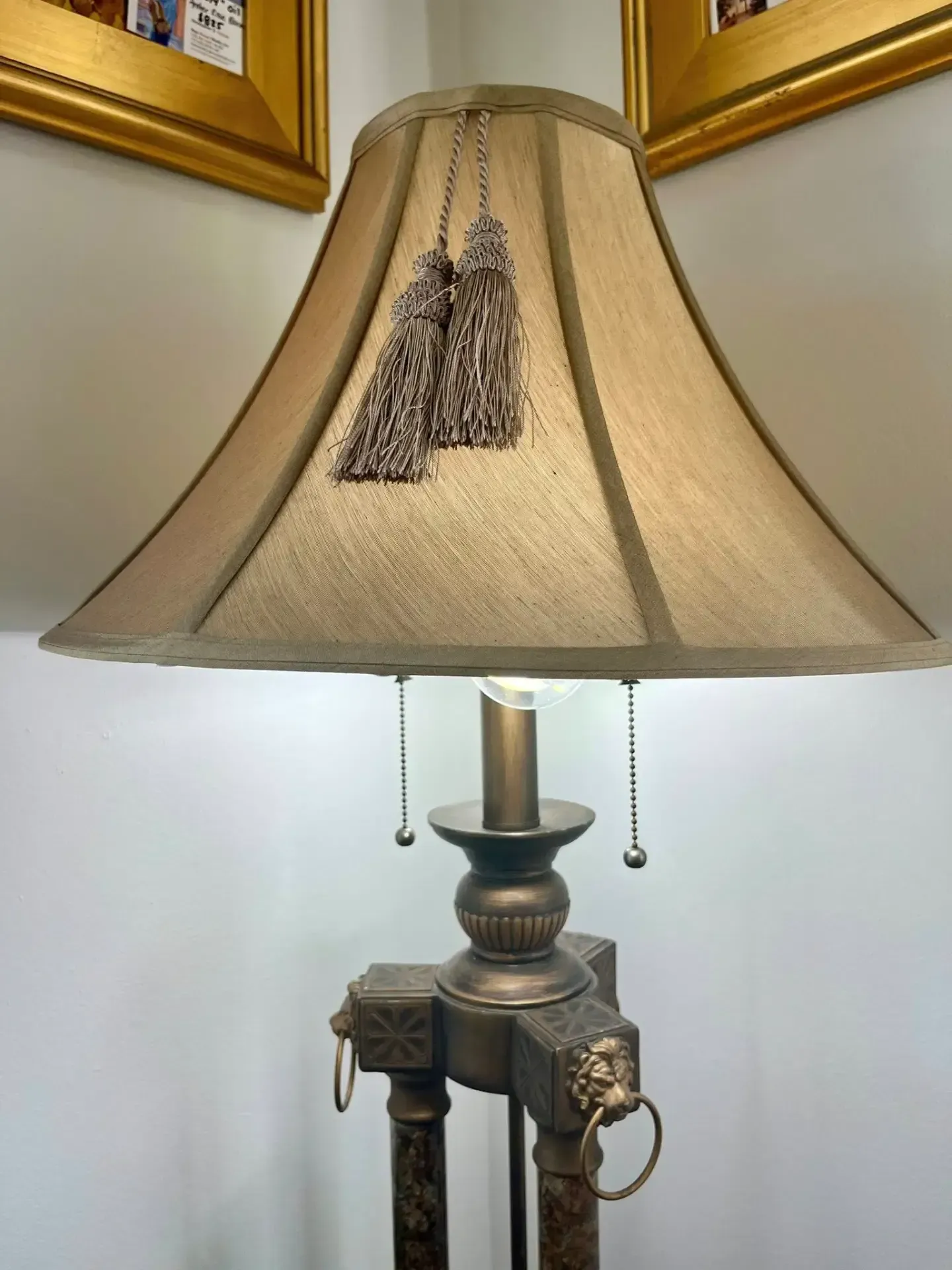 A lamp with a beige fabric shade featuring hanging tassels, set on a bronze base with lion-head accents and ring pulls.