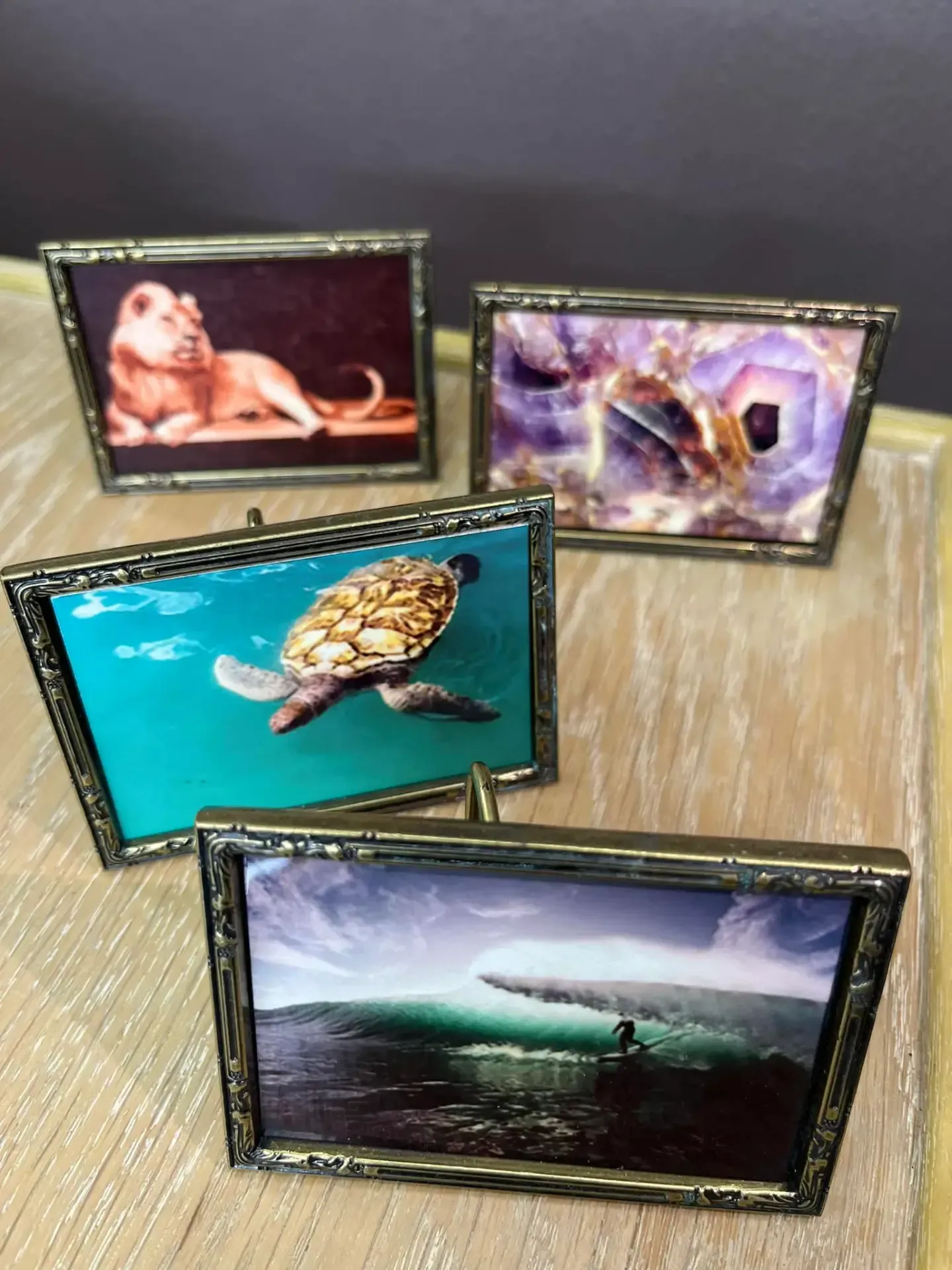 Four small, ornate brass frames on a light wood surface, displaying images of a lion, purple crystals, a turtle, and a wave.