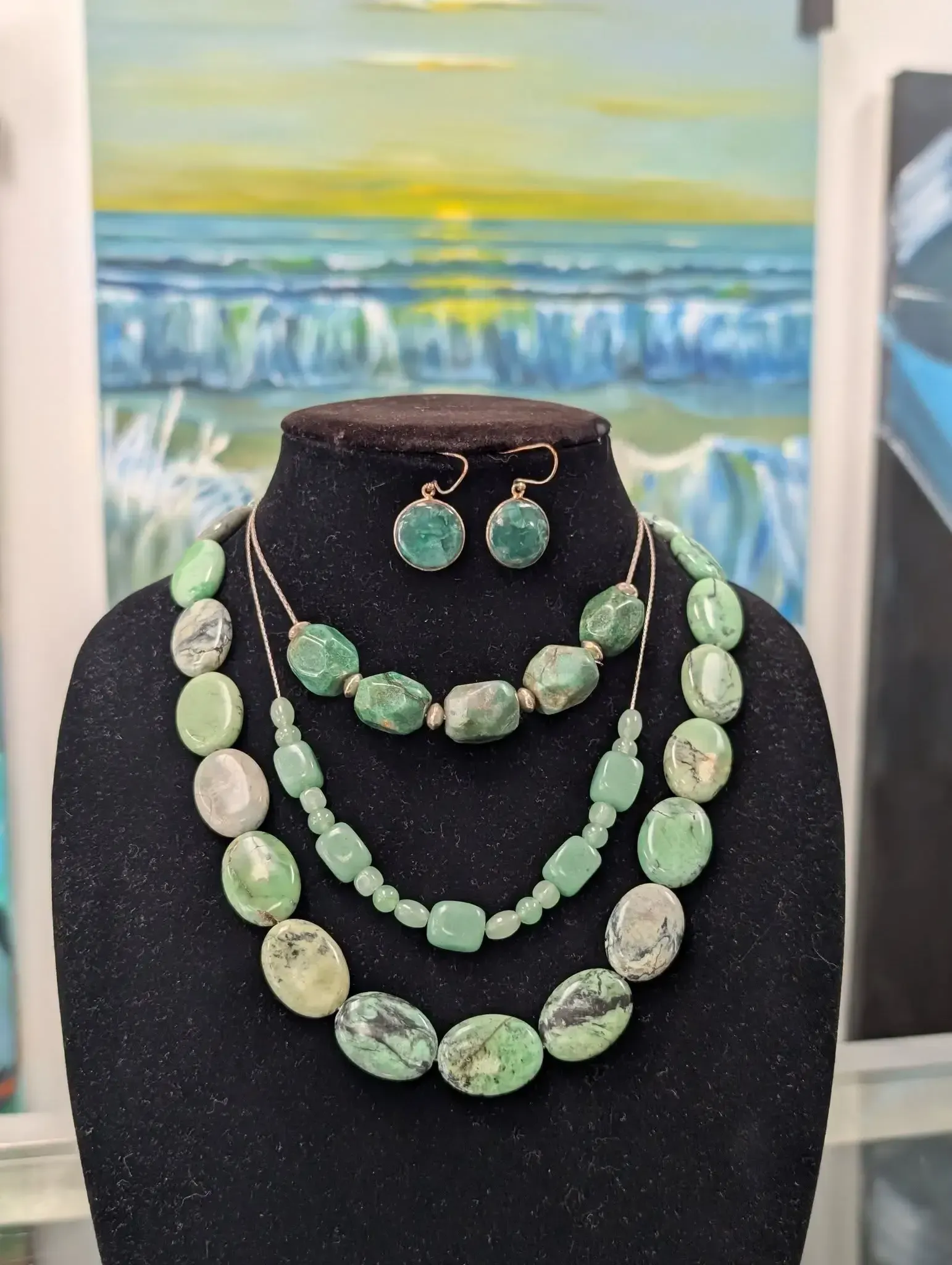 A jewelry set with green, textured oval-shaped stones on a black display bust in front of a painted ocean scene.