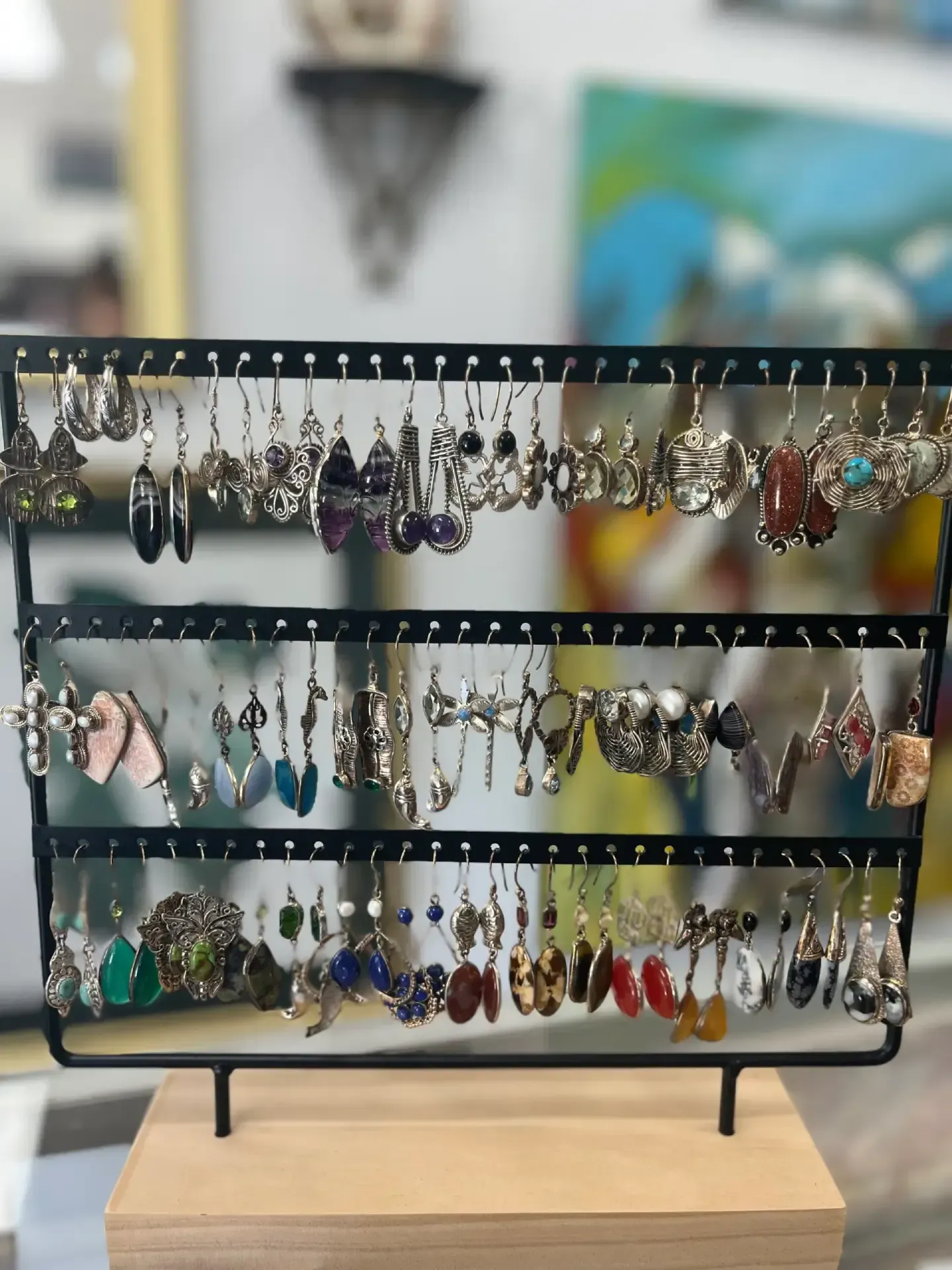 A black three-tiered metal earring display stand holds a diverse collection of dangling earrings on a wooden base.