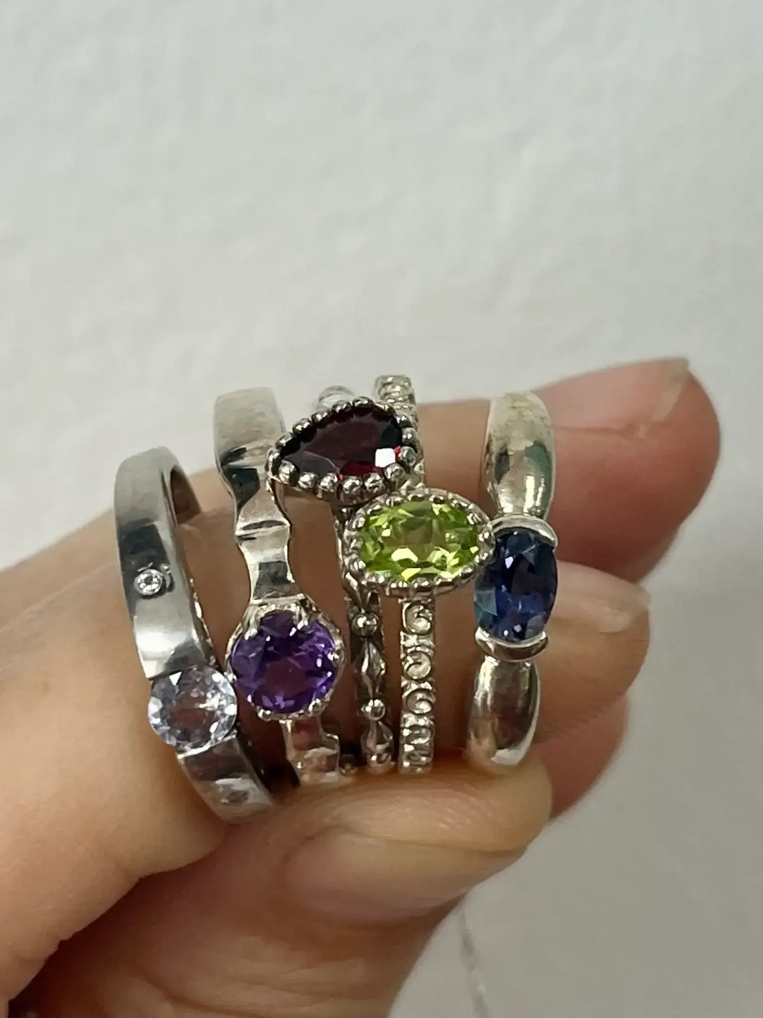 A hand holds five silver rings featuring various gemstones, including purple, red, green, and blue stones.