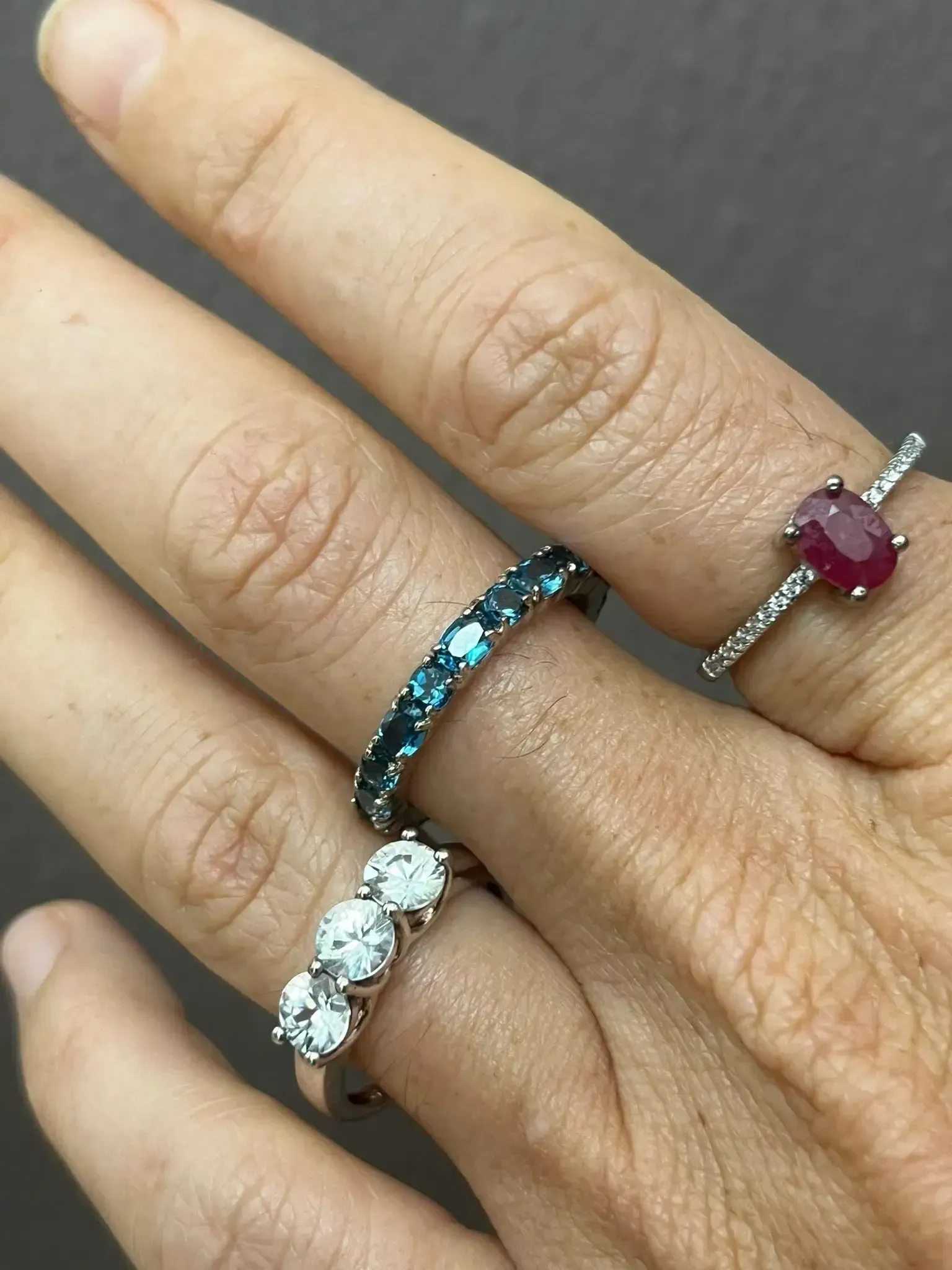 Three rings on a hand: a three-stone diamond ring, a blue gemstone band, and a solitaire ruby ring on the top finger.