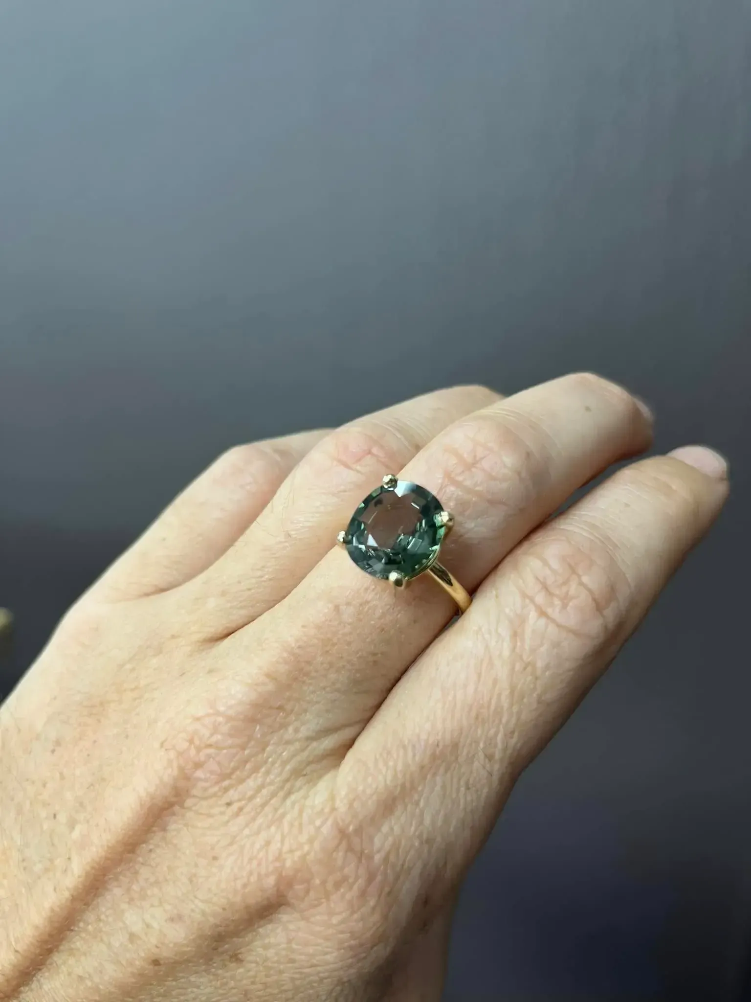 A hand wearing a gold ring featuring a large, square-cut green gemstone against a solid grey background.