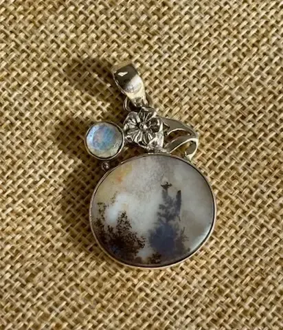 A silver pendant featuring a round dendritic agate with tree-like inclusions and a small, round moonstone accent.