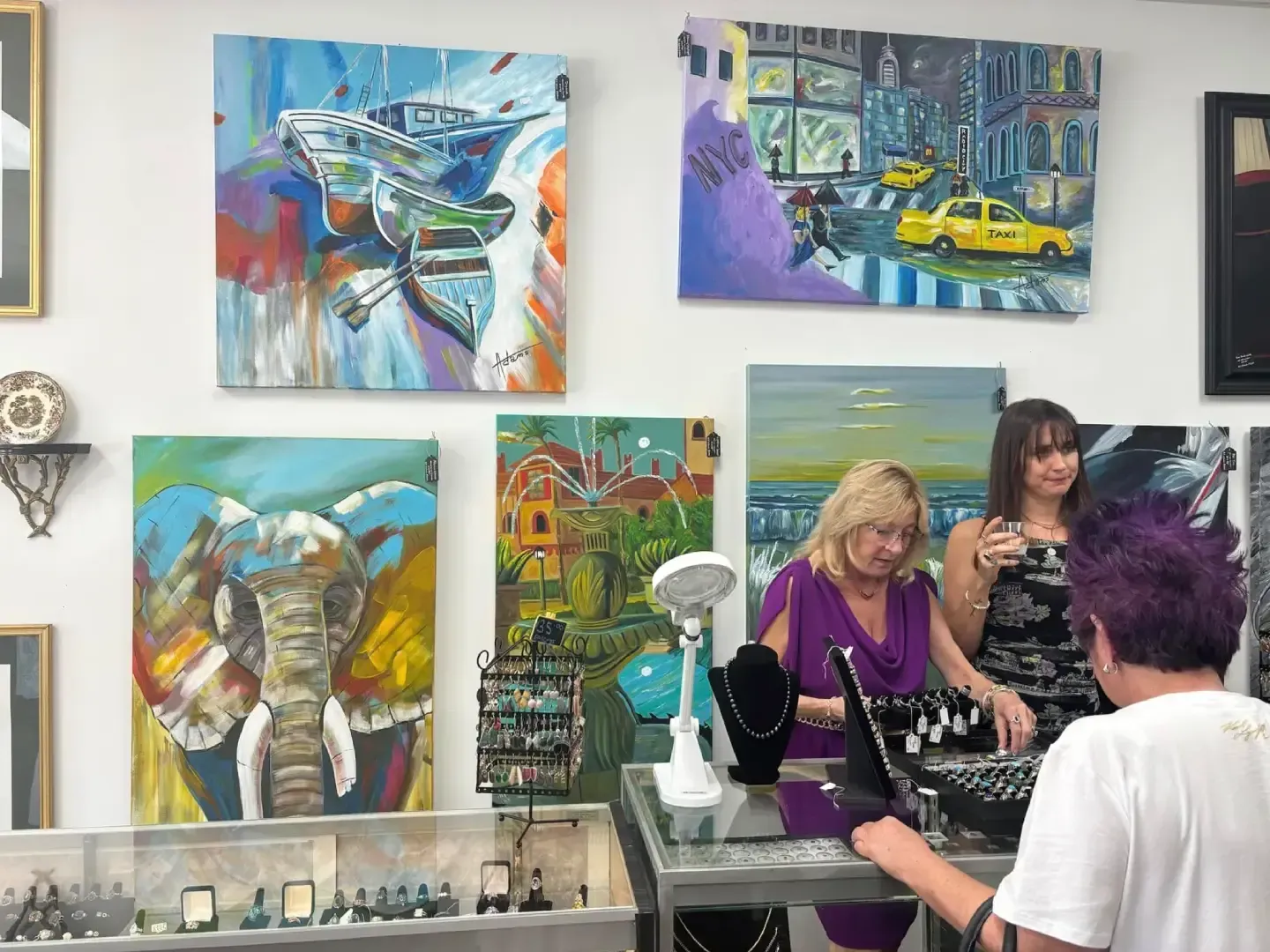 Two people in an art gallery, looking at jewelry on a counter beneath several colorful paintings displayed on the wall.