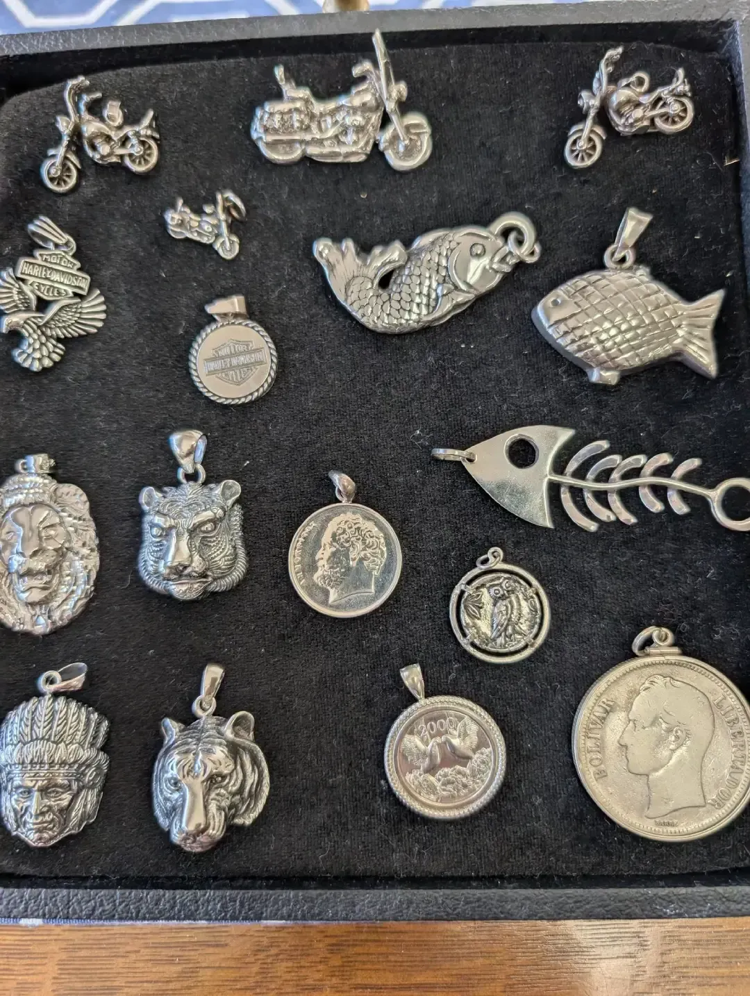 A display tray containing various metallic silver-toned pendants, including motorcycles, animals, and historical coins.