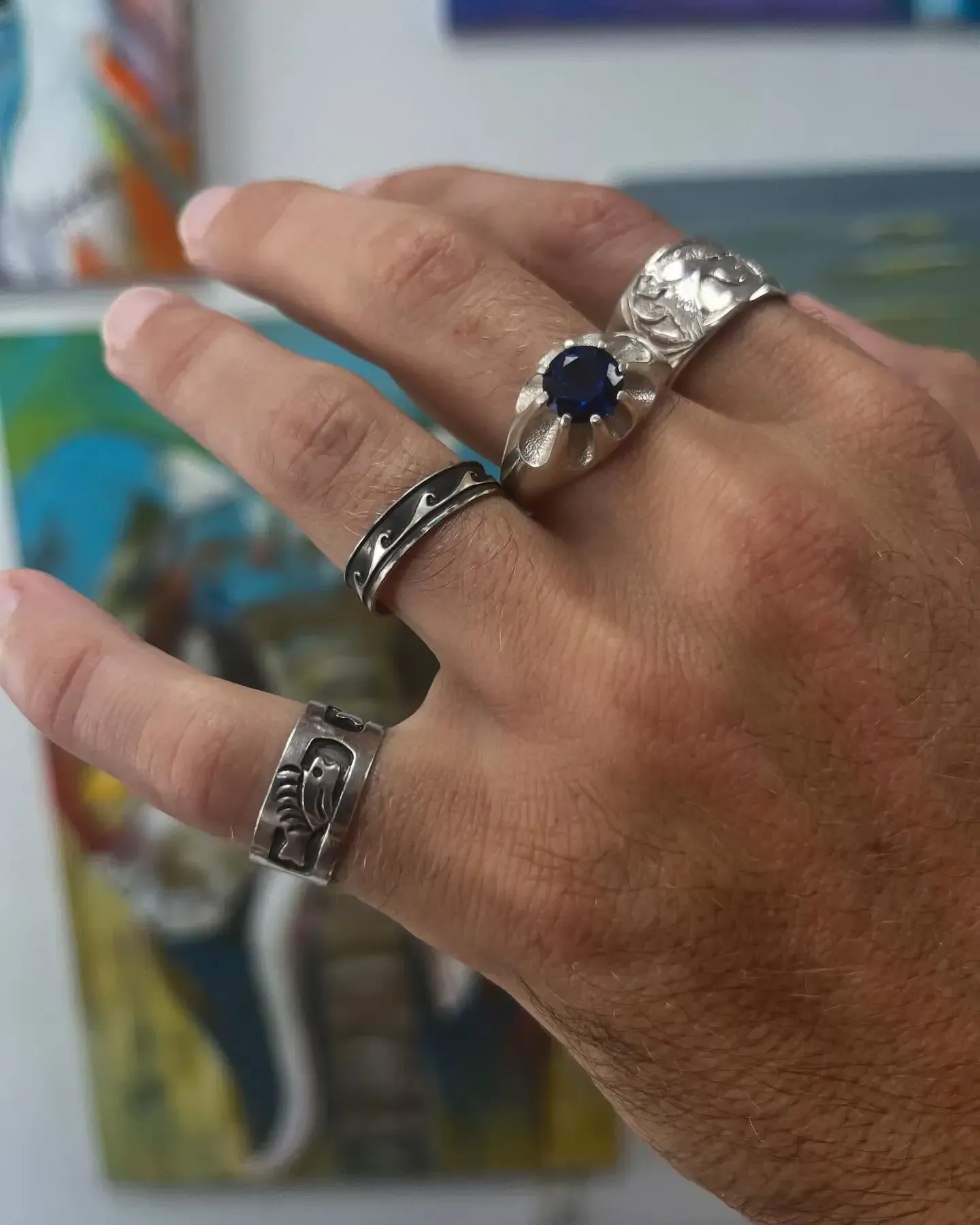 A hand wearing four distinct silver rings, including one with a dark blue stone, against a colorful, blurry background.