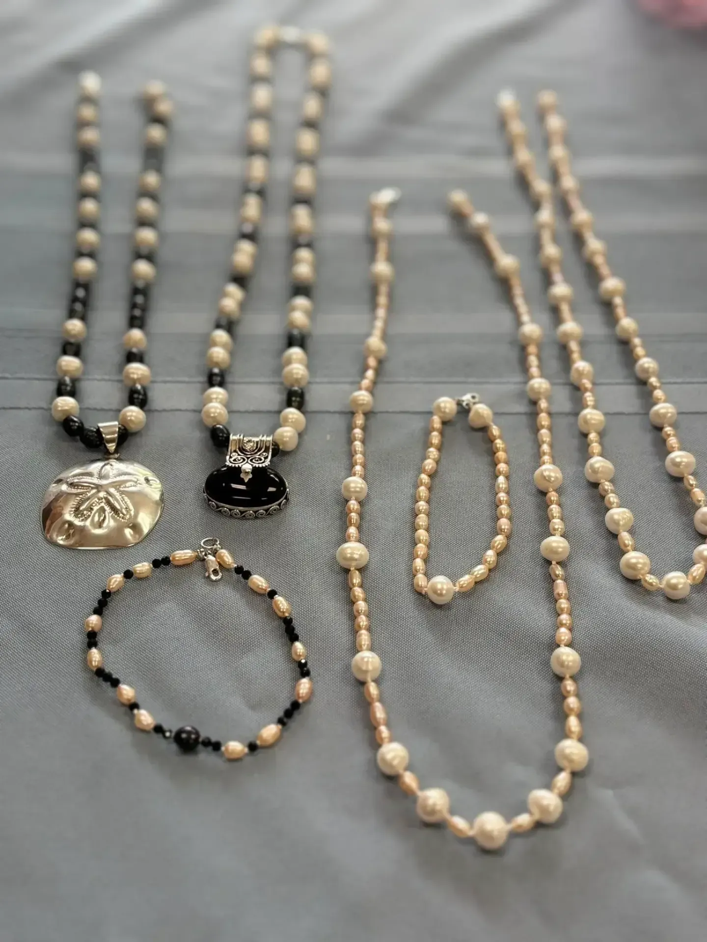 A collection of beaded necklaces and bracelets featuring white pearls, black beads, and silver pendants on a gray cloth.