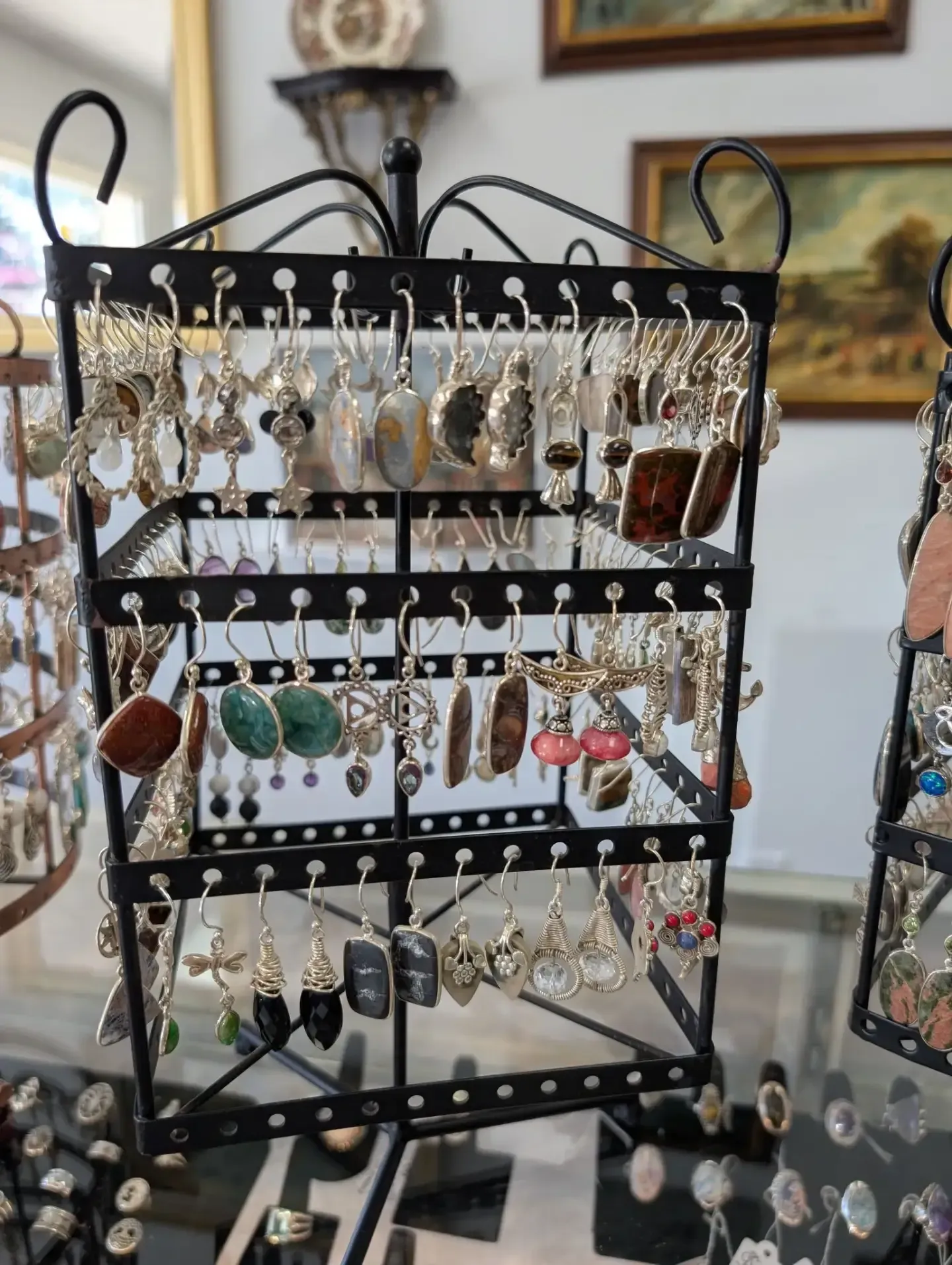 A black metal display stand holds rows of various hanging earrings, featuring diverse shapes, colors, and stone designs.