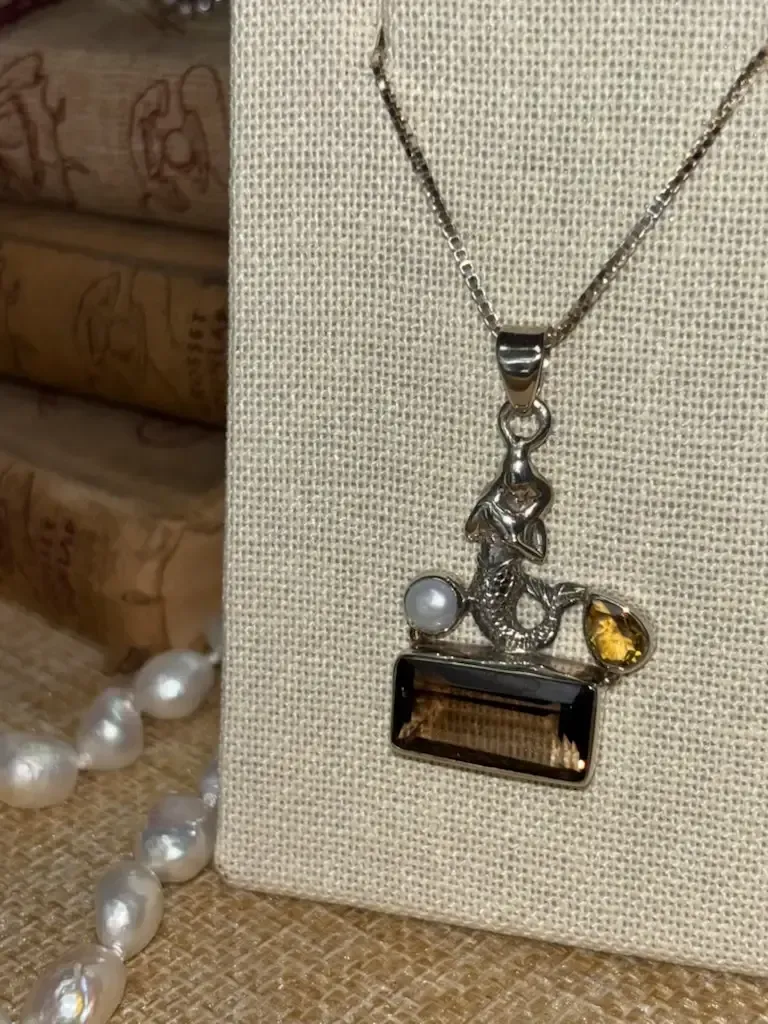 A silver mermaid pendant with a white pearl, yellow gemstone, and large rectangular brown stone on a fabric display.