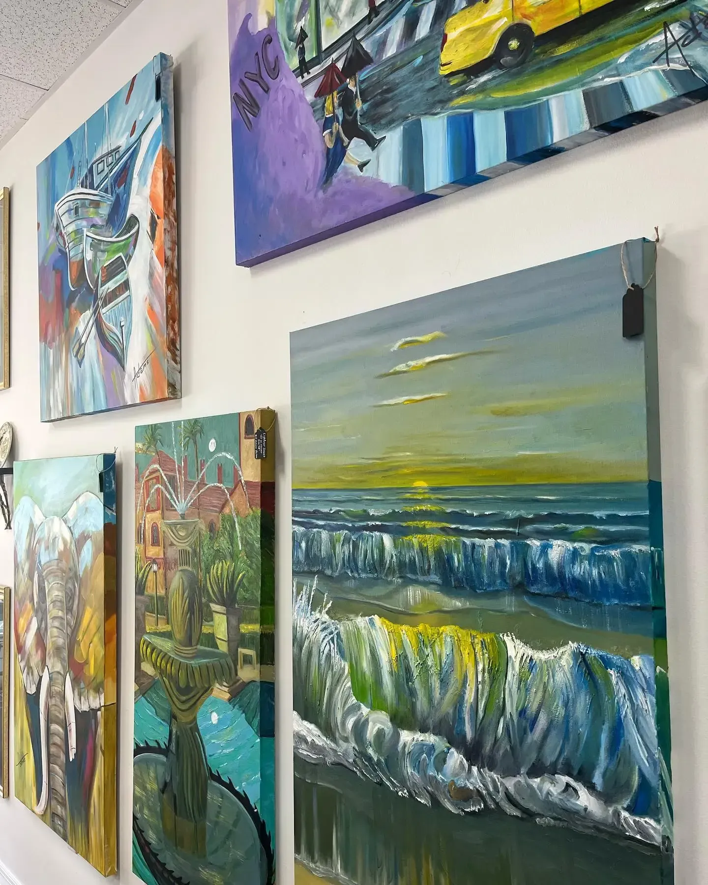 Five diverse landscape and abstract paintings hung on a white wall in an art gallery.