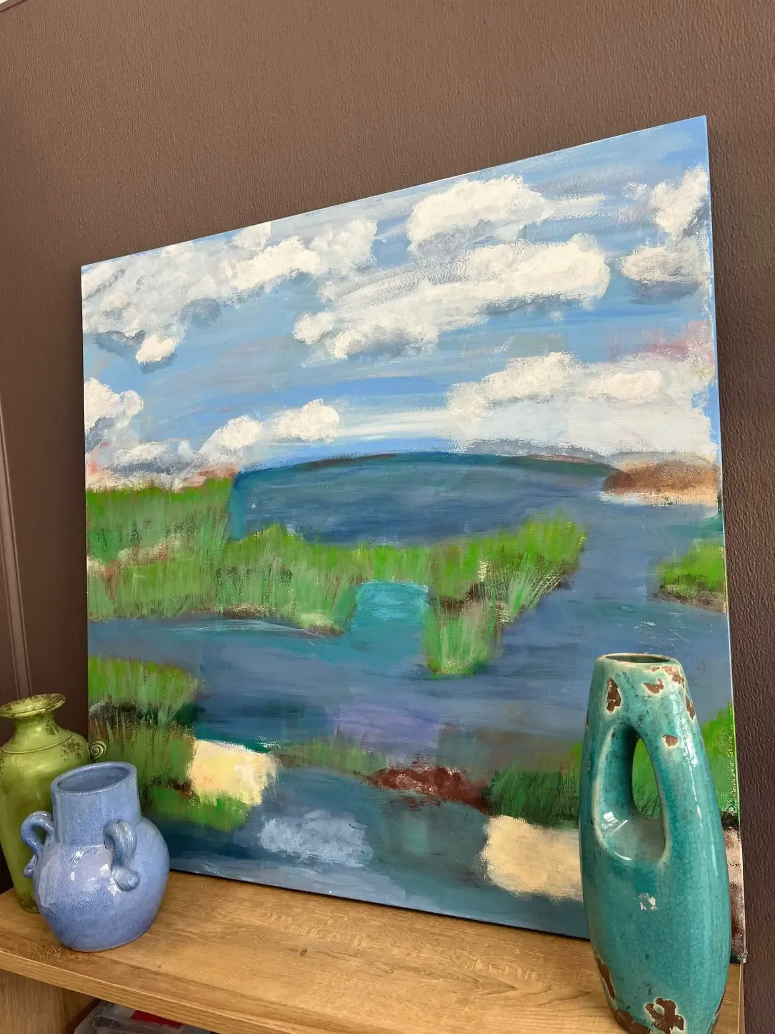An oil painting of a blue, grassy landscape under a cloudy sky sits on a shelf next to three ceramic vases.