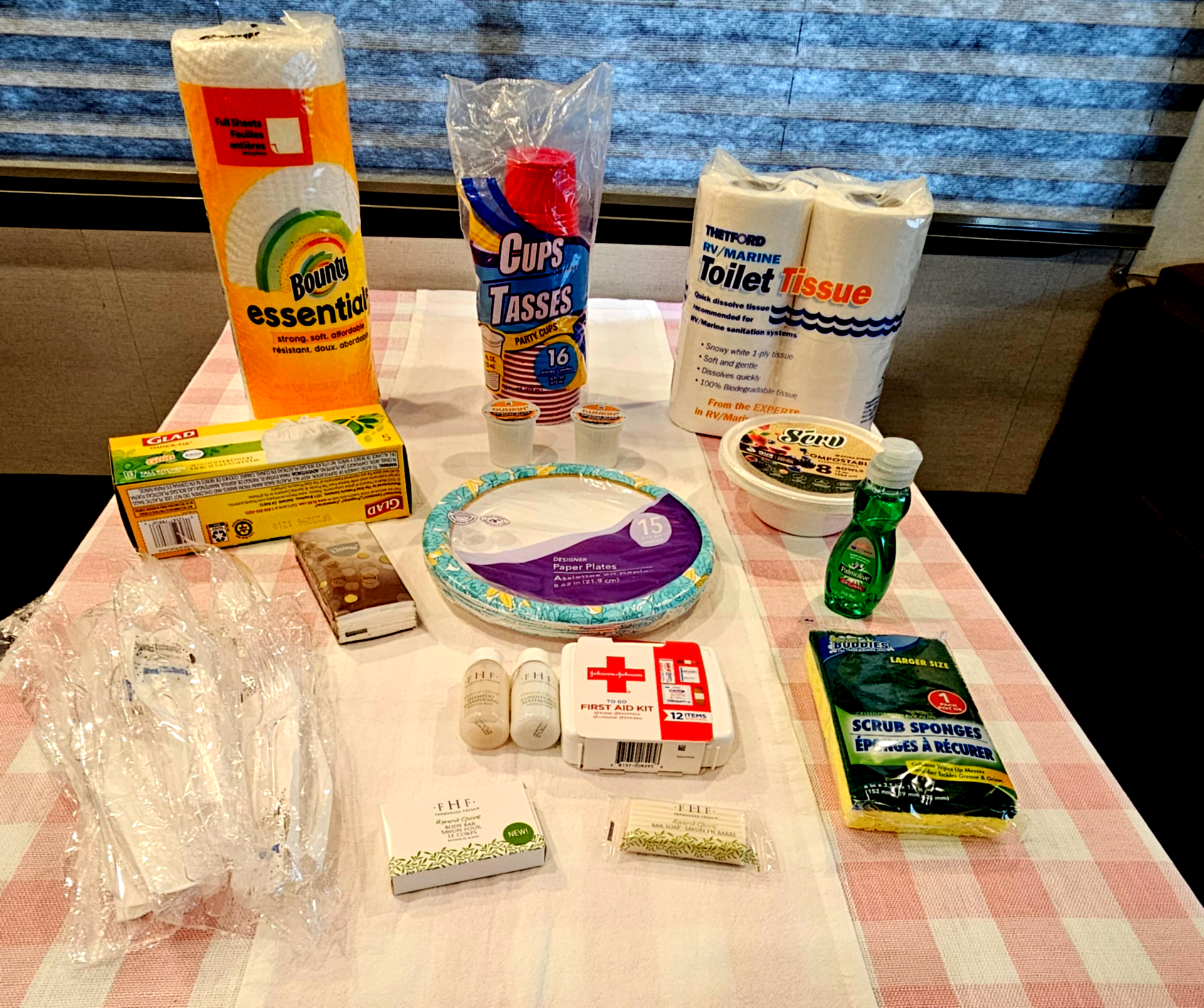 A table with toilet paper, paper towels, plastic kitchen utensils, paper plates, tissues, plastic cups, dish soap, body soaps and shampoo.