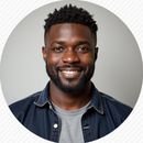 Smiling Black man with a beard, wearing a denim shirt and grey t-shirt, against a light grey background.
