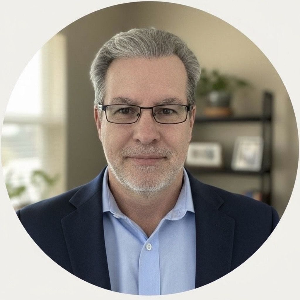 Man with graying hair and glasses wearing a light blue shirt, indoors, smiling slightly.