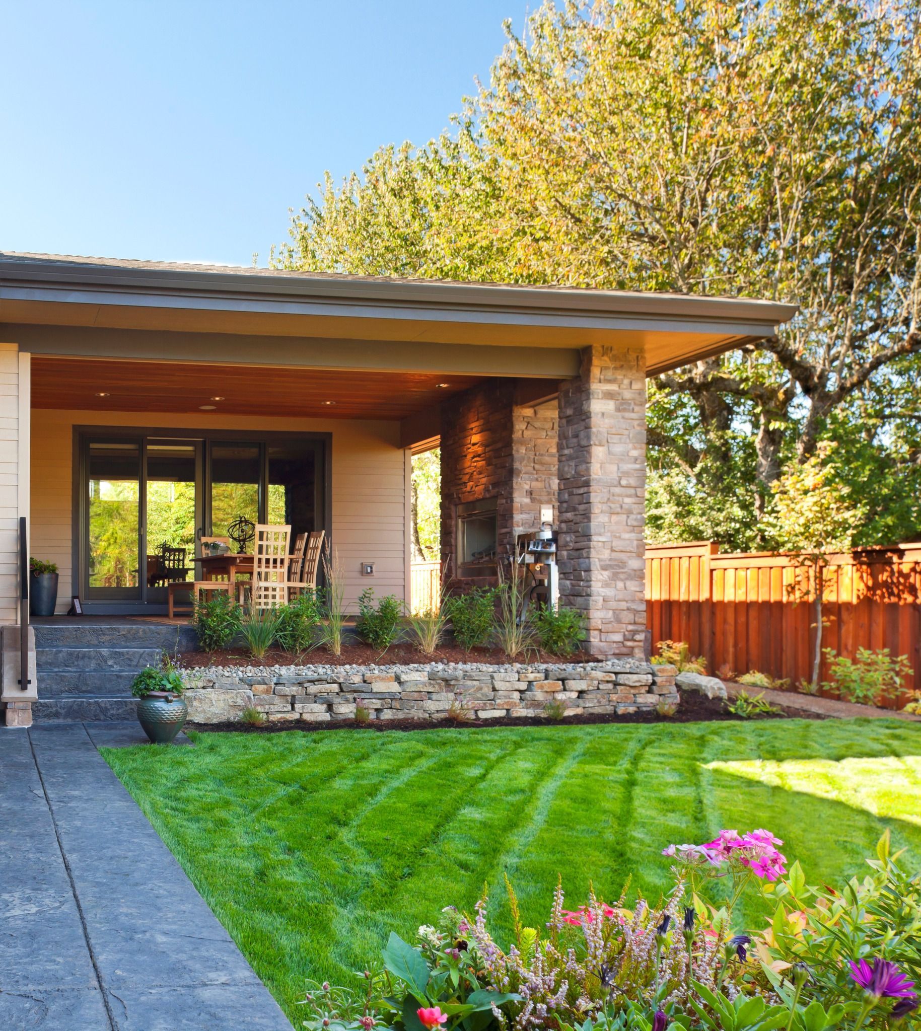 Backyard patio with green lawn, flower bed, stone wall, and covered seating area.
