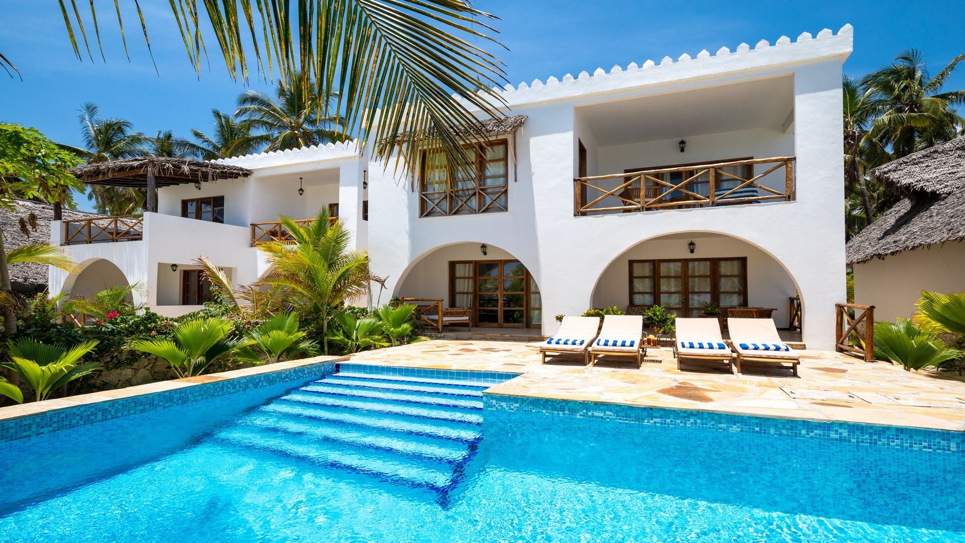White beachfront villa with a swimming pool, sun loungers, and palm trees under a blue sky.