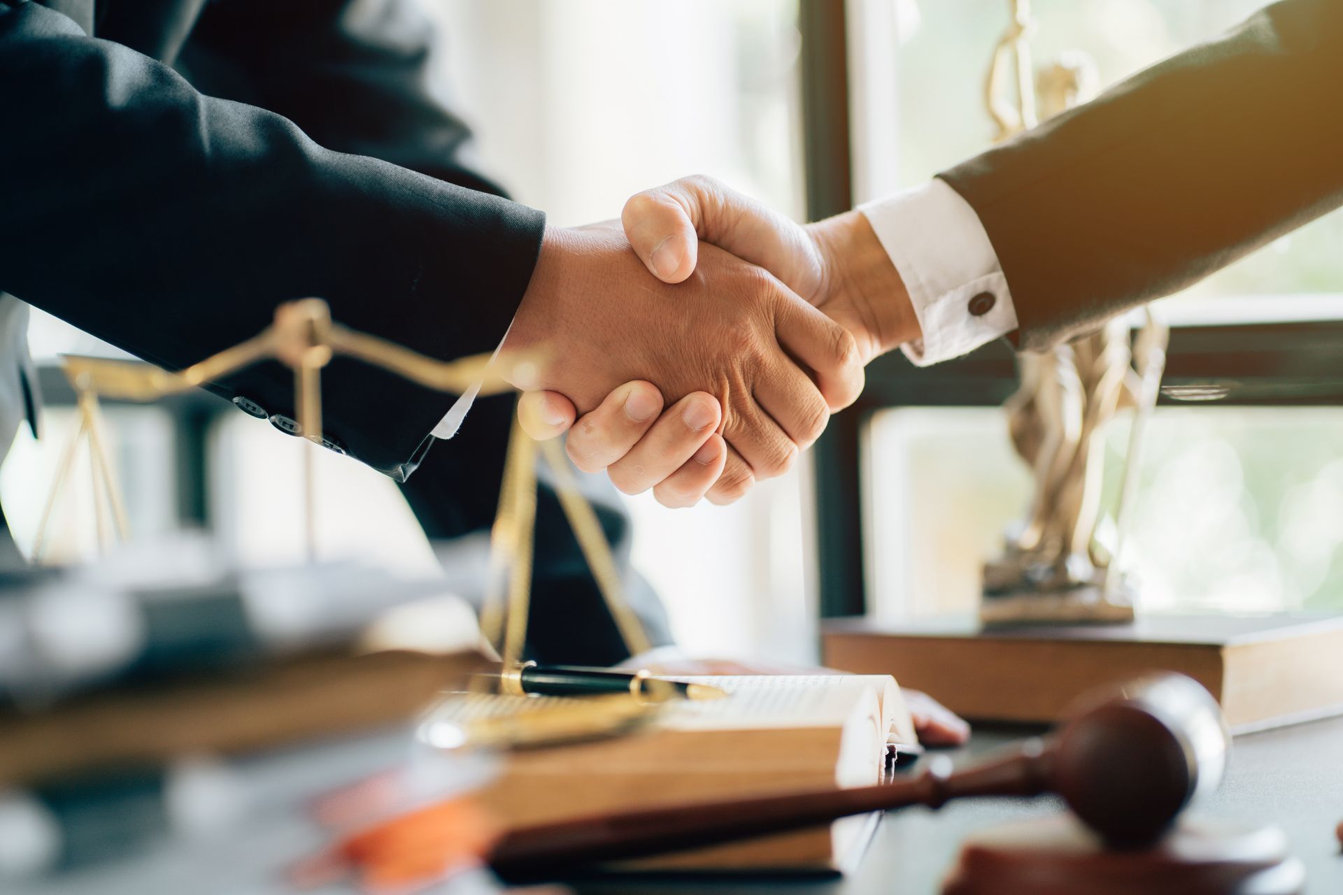 Two professionals in business attire shake hands over a desk containing a legal scale and a wooden gavel.