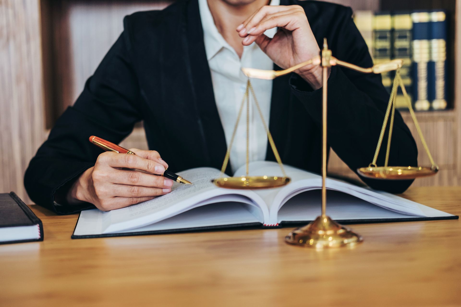 Lawyer writing notes with scales of justice on desk in law office with legal books background.