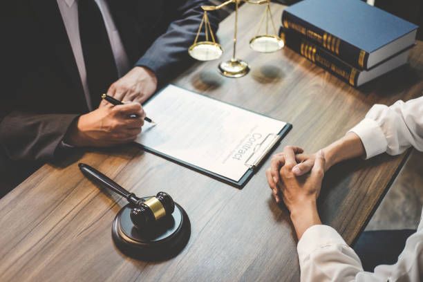 Lawyer consulting client at desk with contract, gavel, scales and law books in office meeting.