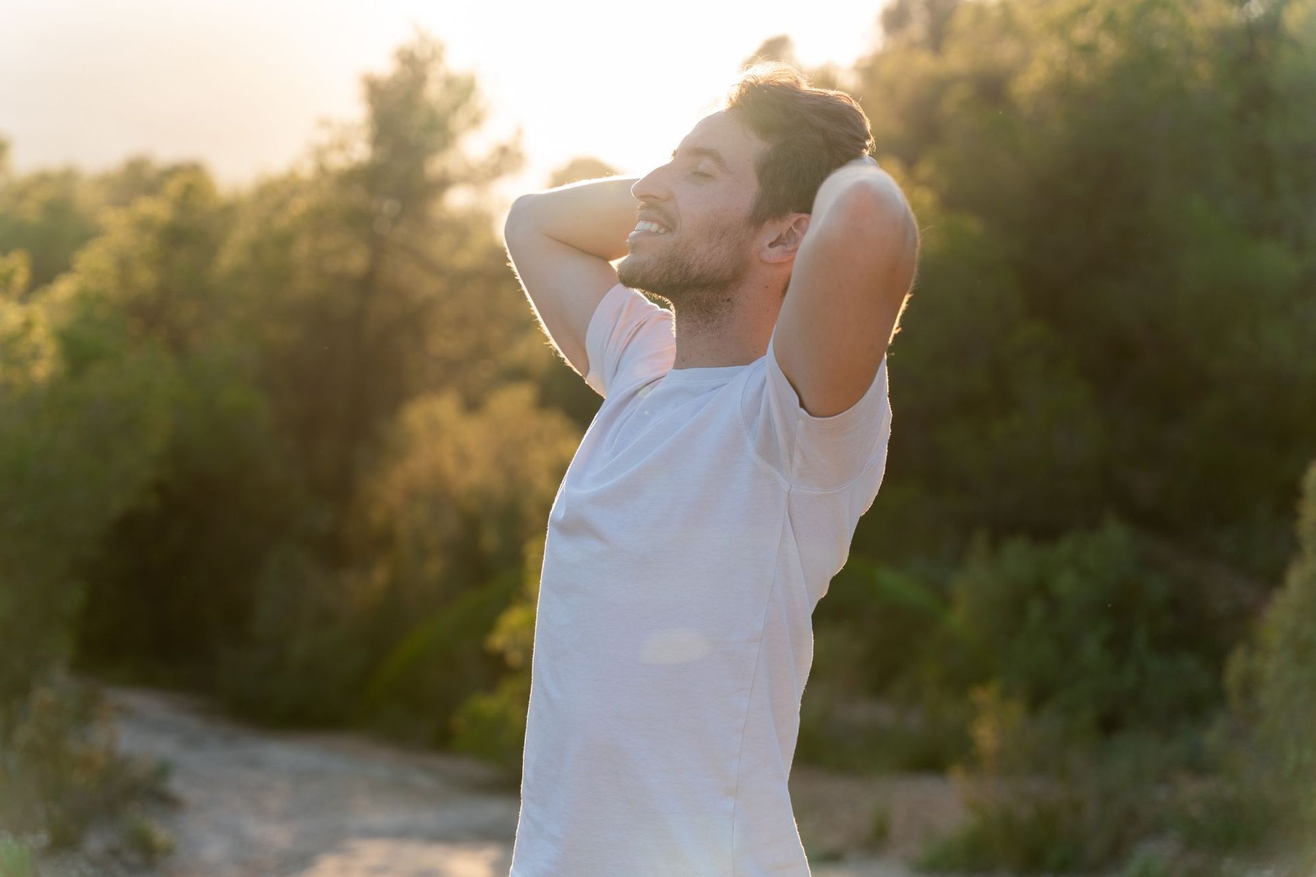 A man in a white shirt is standing in the woods with his hands behind his head.