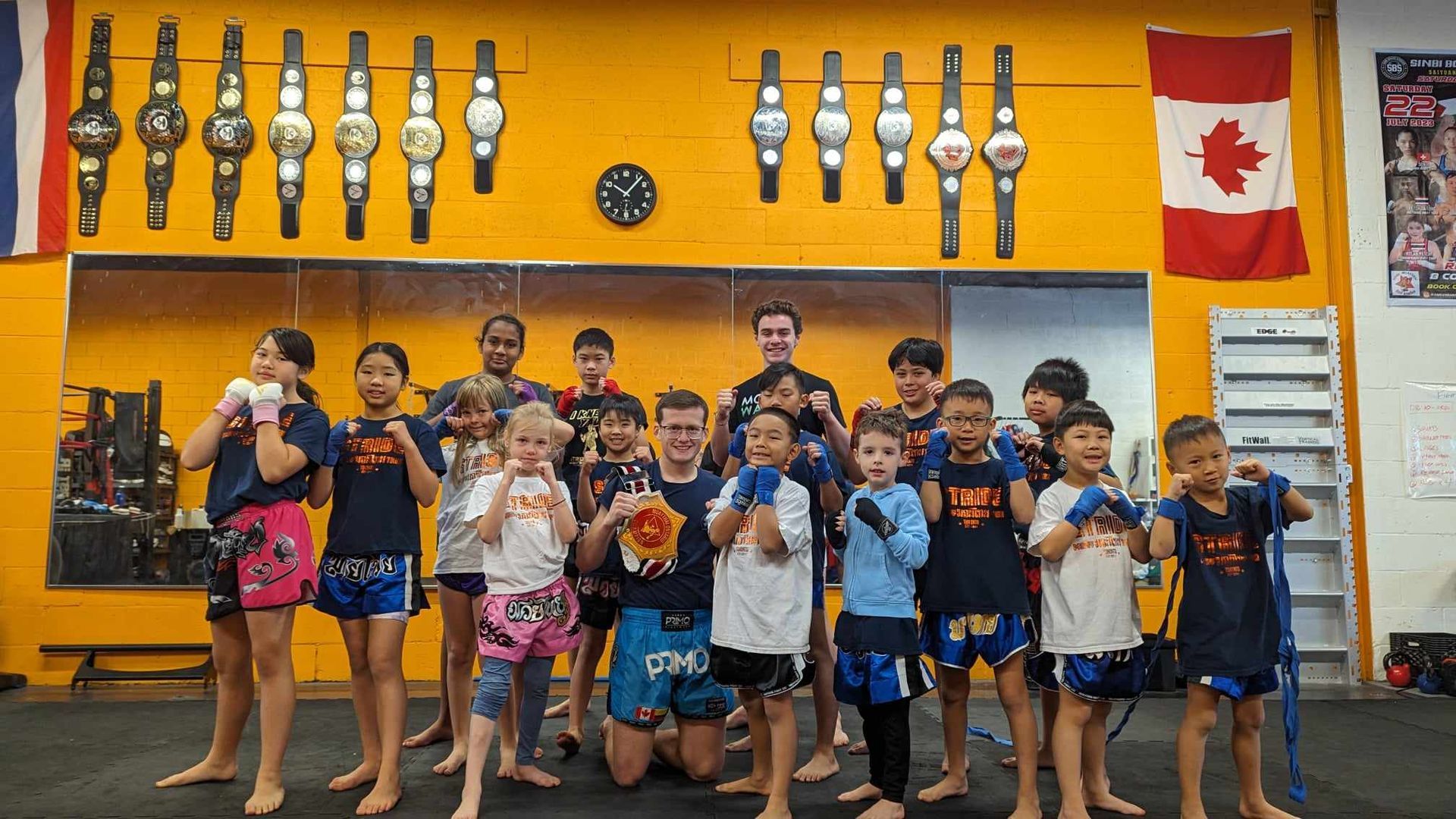 Group of kids and adults in Muay Thai gear, posing in a gym with championship belts on the wall and a Canadian flag.