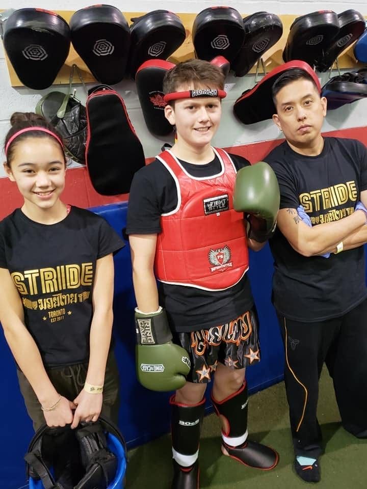 Three people in a gym: a young boy in boxing gear smiles with two others wearing “STRIDE” shirts.