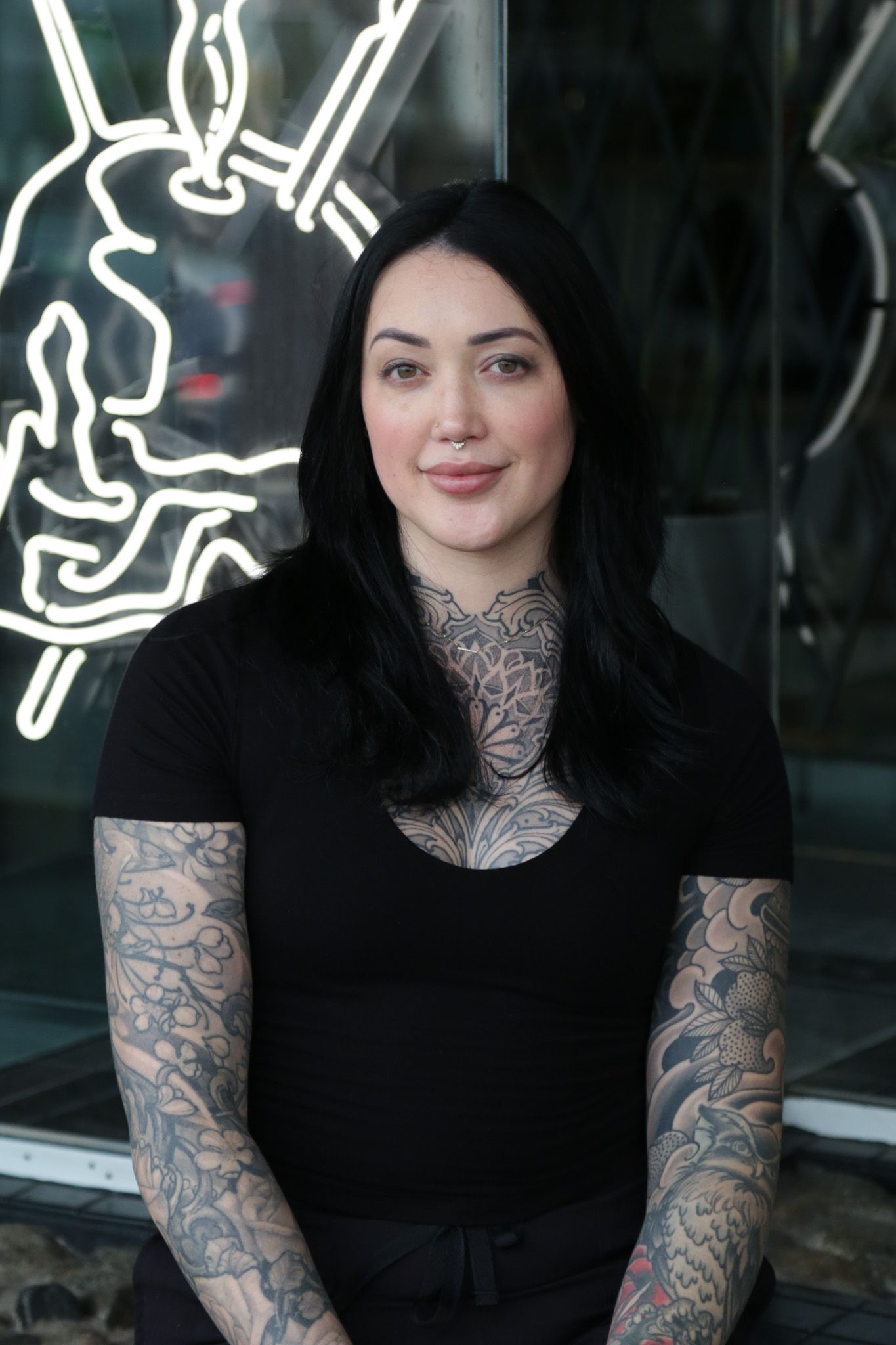 Woman with tattoos stands in front of a neon sign.
