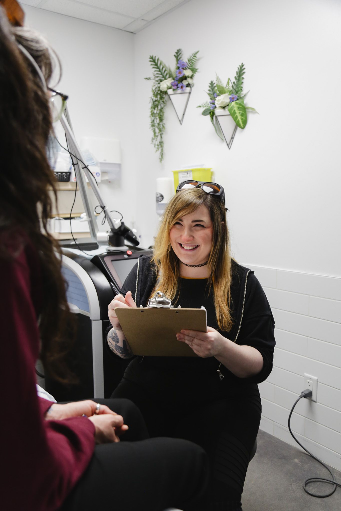 Woman in black outfit, smiling, holding clipboard, talking to another woman in a medical office setting.