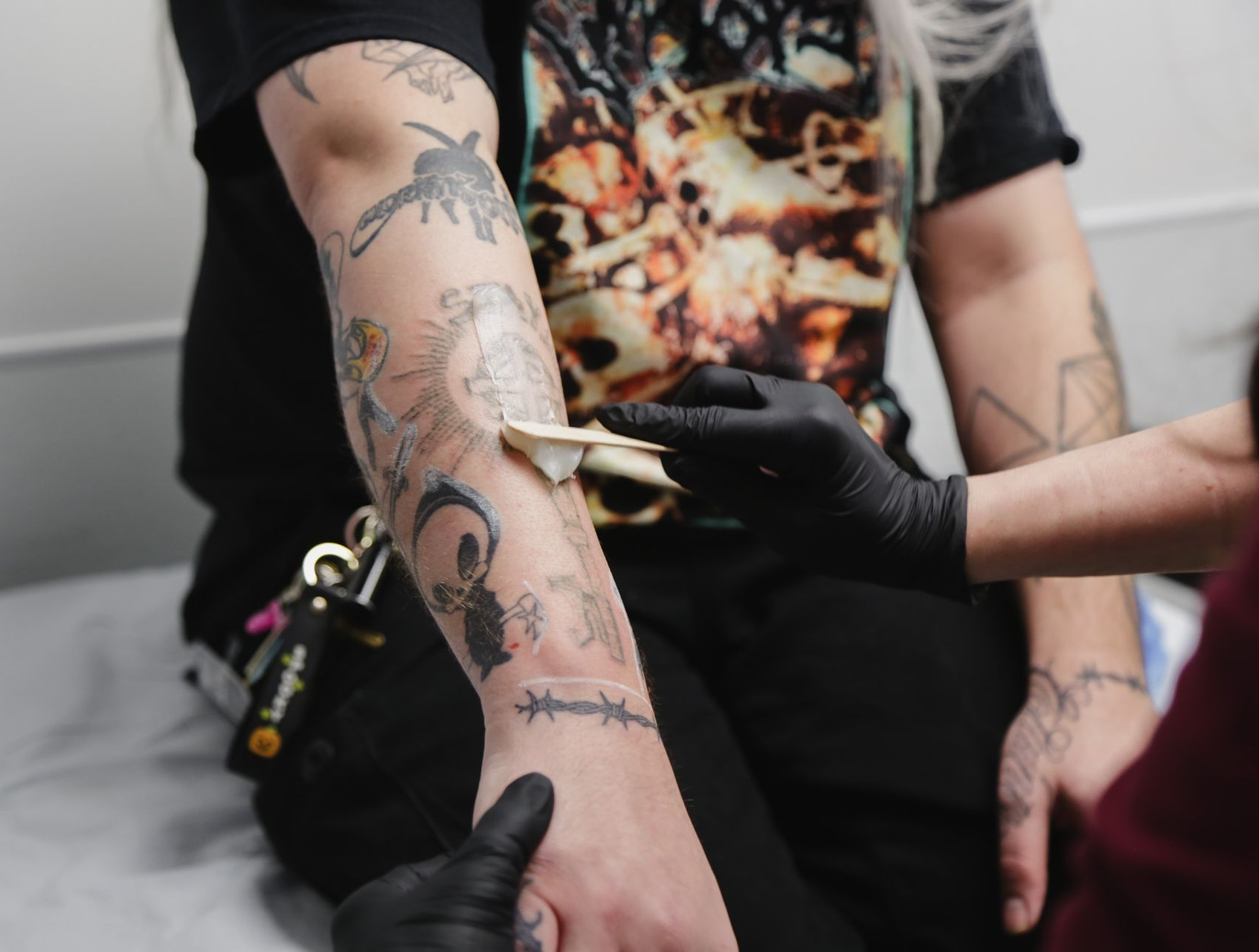 Person getting tattoo on arm; artist wearing gloves applies cream to the artwork.