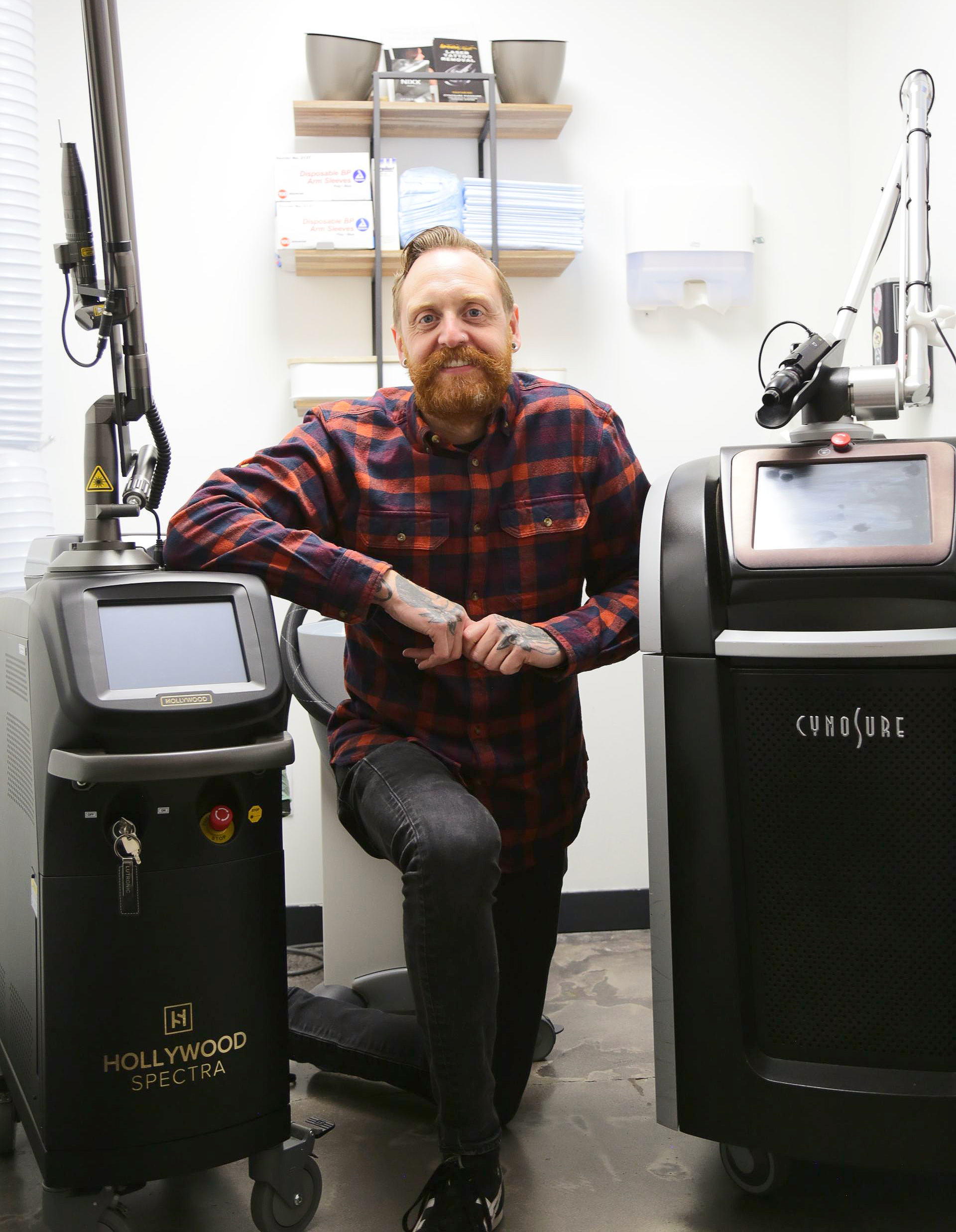 Man with red beard poses between two laser machines. He wears plaid shirt in medical setting.