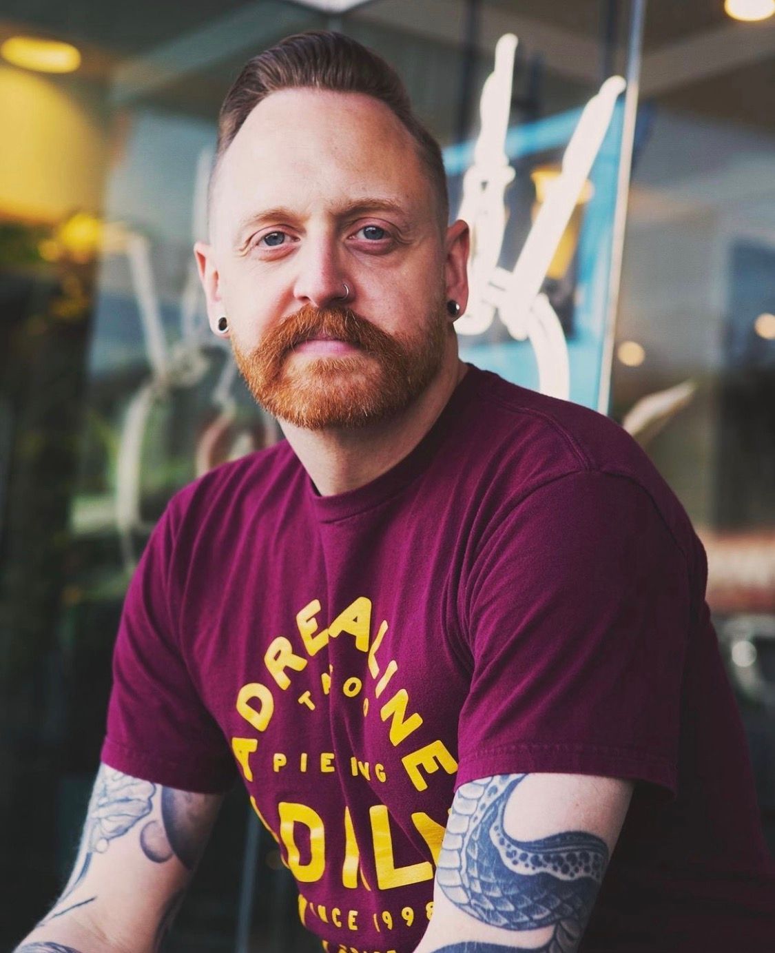 Man with red hair, mustache, tattoos, and burgundy shirt; sitting by a window.
