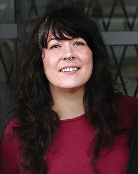 Woman with dark hair smiles, wearing a red shirt, nose ring. She is near a glass window.