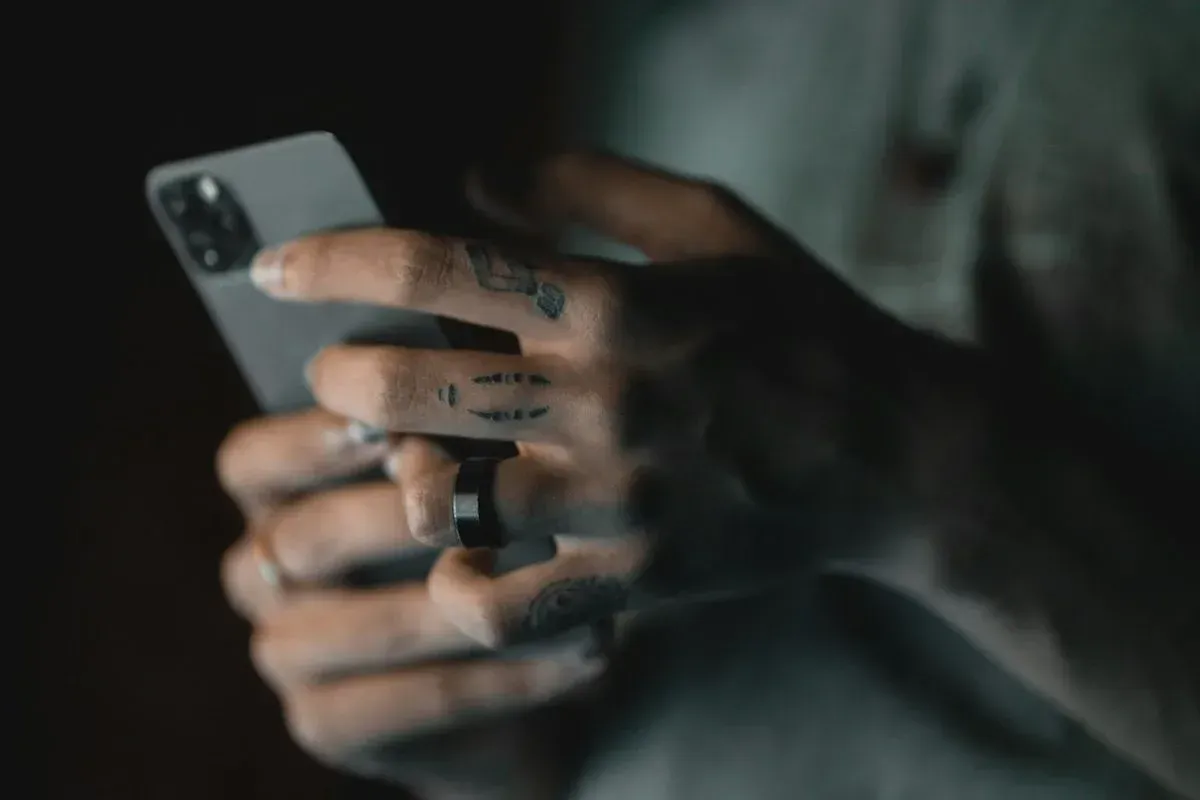 Close-up of tattooed fingers holding a smartphone in a dimly lit setting.